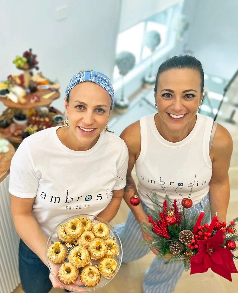 Two smiling women in white 'Ambrosia' t-shirts, one holding a plate of assorted donuts and the other holding a festive floral arrangement with red decorations, standing in a bright, modern room with a table of food in the background.