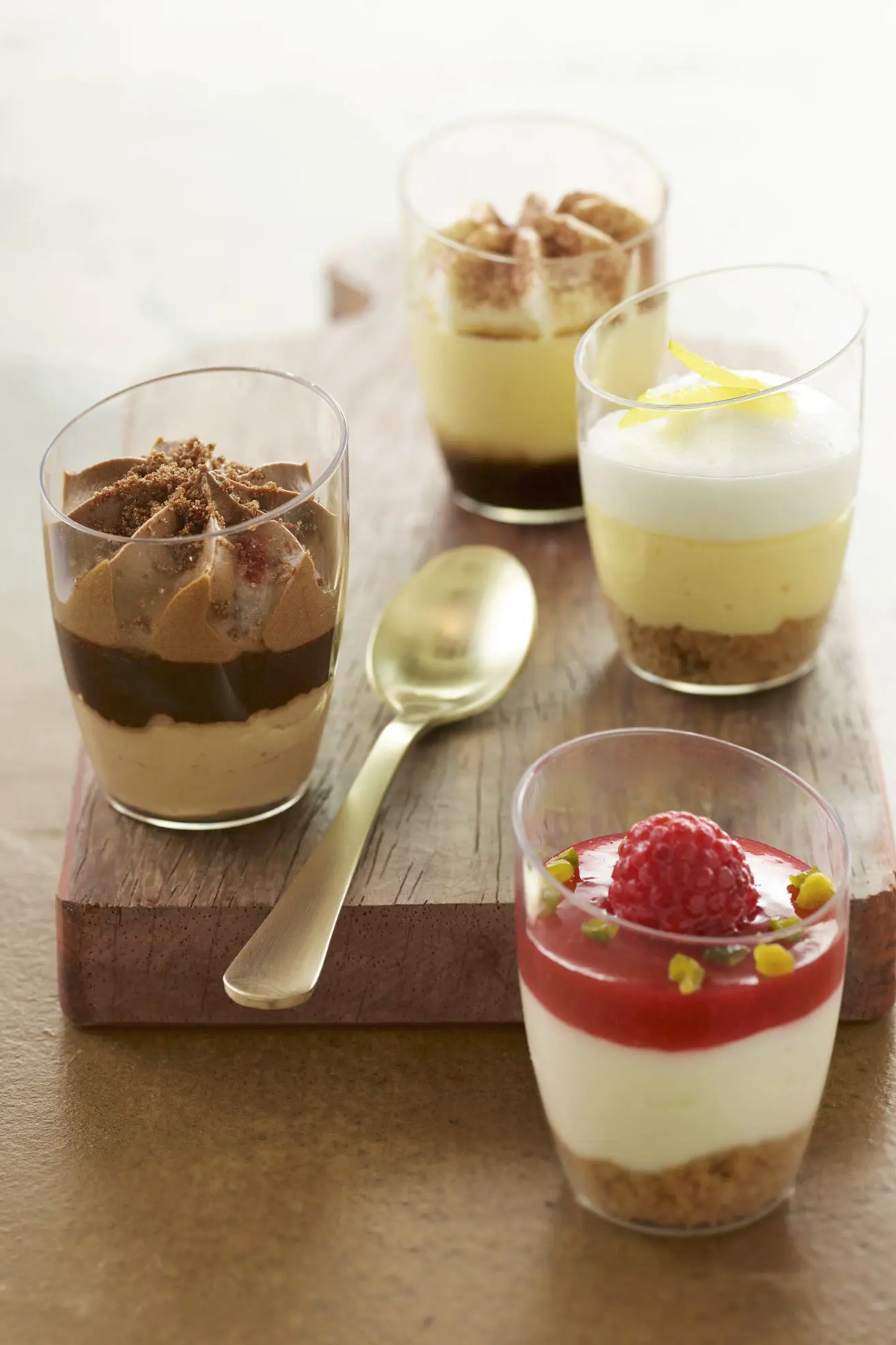 Four individual layered desserts in clear glasses with different toppings, placed on a wooden board and a beige surface.
