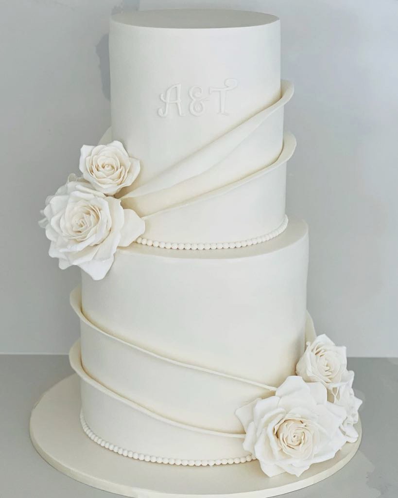 Elegant white tiered wedding cake decorated with white roses and delicate piping, with initials 'A & T' on the top tier.