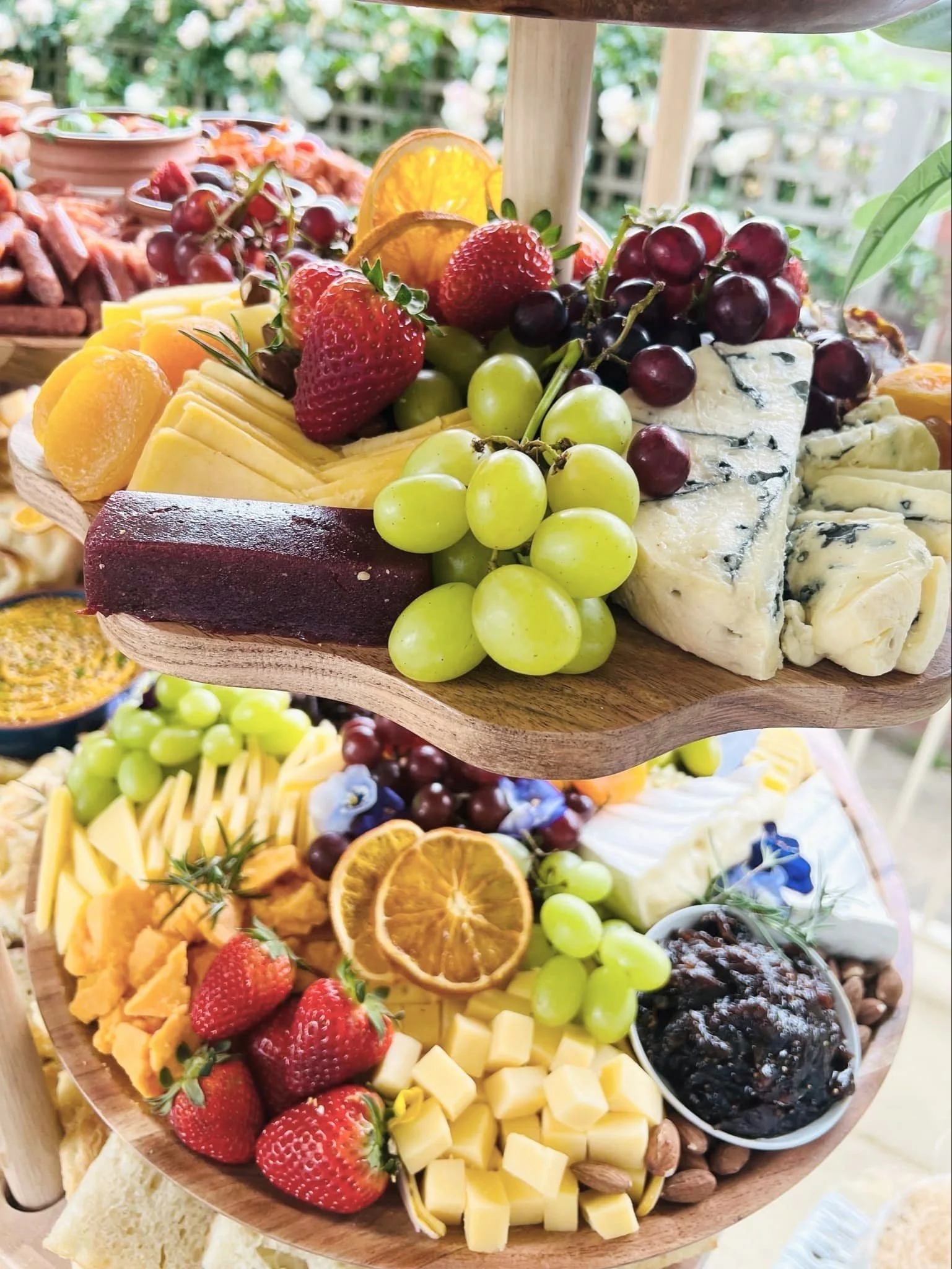 Two-tiered wooden cheese and fruit platter with strawberries, grapes, blue cheese, cheddar, dried orange slices, and fruit preserves.