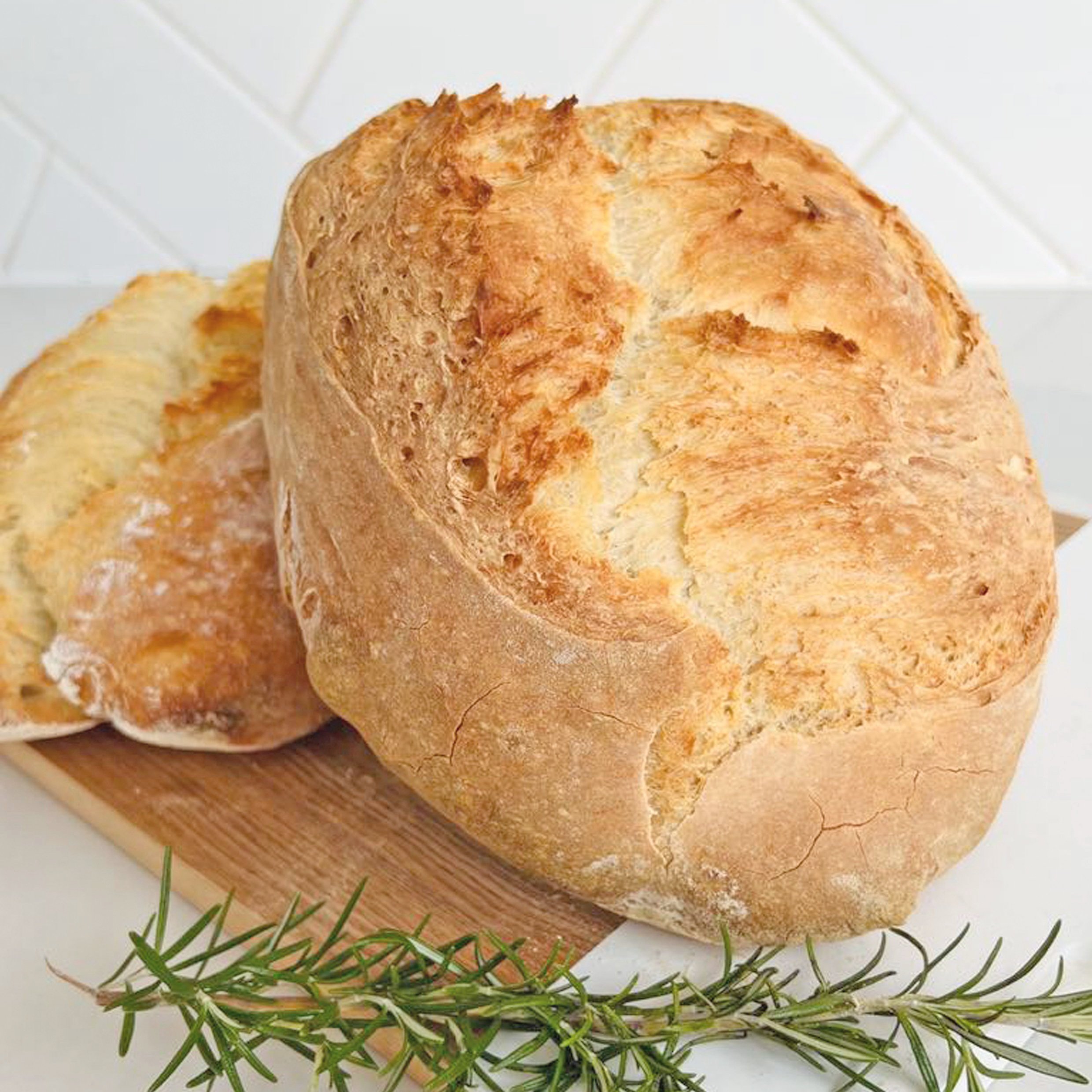 Freshly baked loaf of bread on a wooden cutting board with a sprig of rosemary in the foreground.