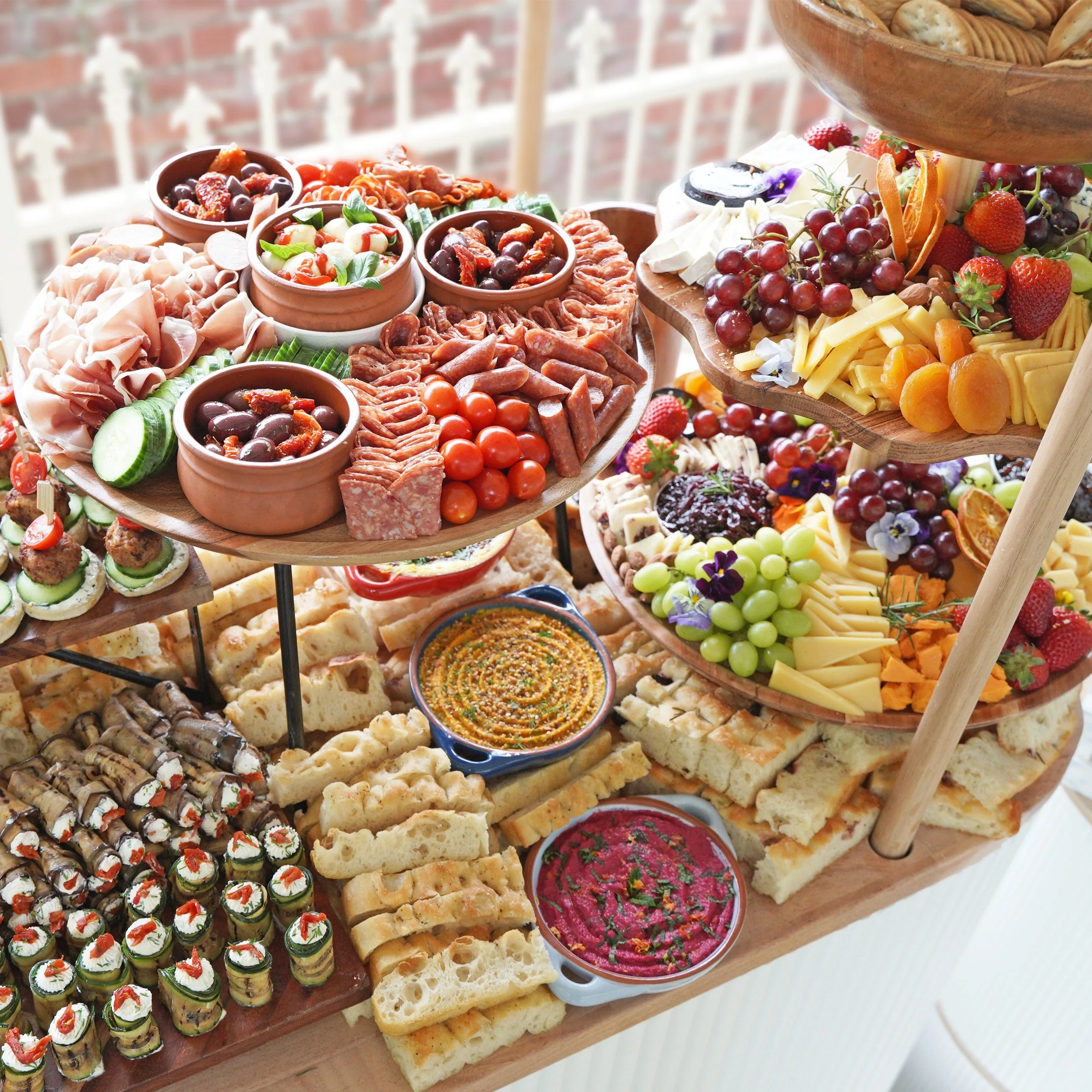 A wooden serving tray with various appetizers including cured meats, cherry tomatoes, olives, cheese, fruits, bread, and dips. Some appetizer cups contain salads and vegetables, with other items like rolled meats, sliced bread, and decorated mini sandwiches.