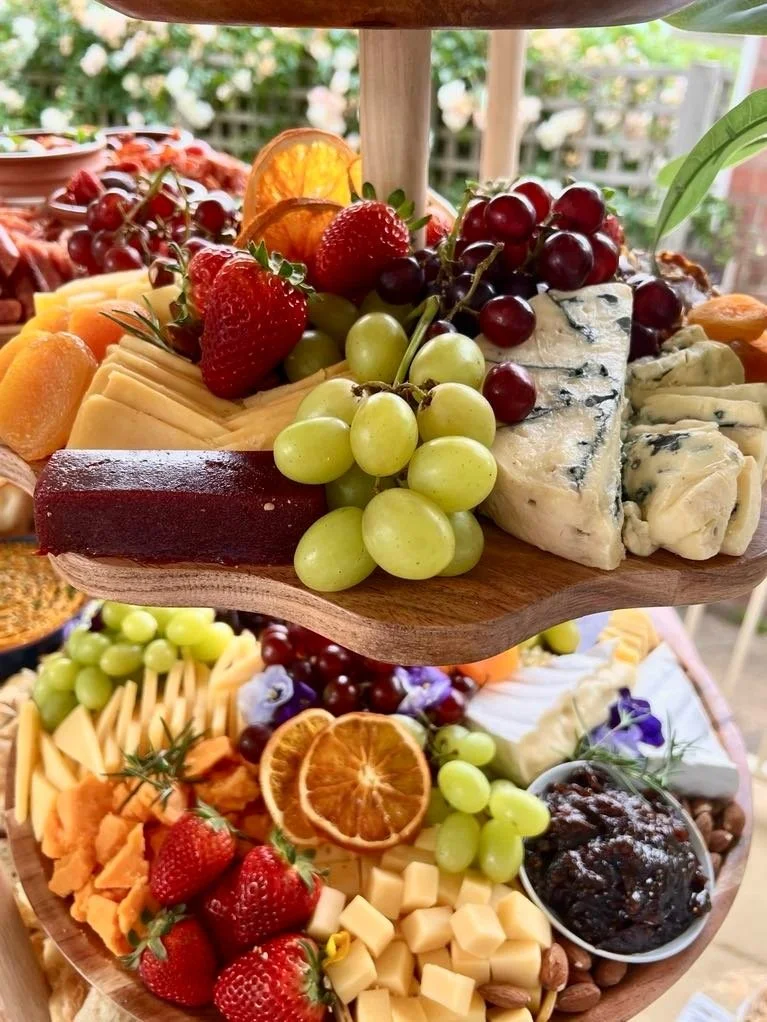 Two-tiered wooden cheese and fruit platter with grapes, strawberries, cheese, dried orange slices, blue cheese, and chutney in a garden setting.