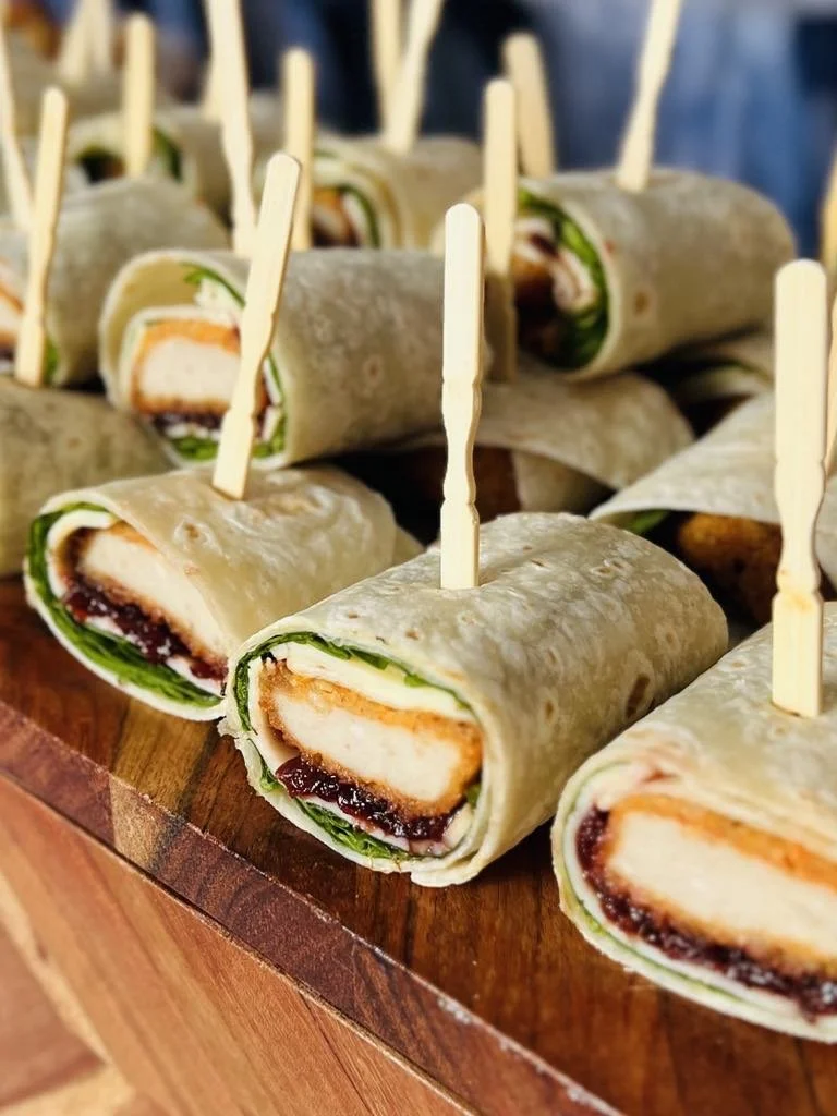 Close-up of mini sandwich wraps with chicken, lettuce, and sauce, secured with small wooden skewers, arranged on a wooden surface.