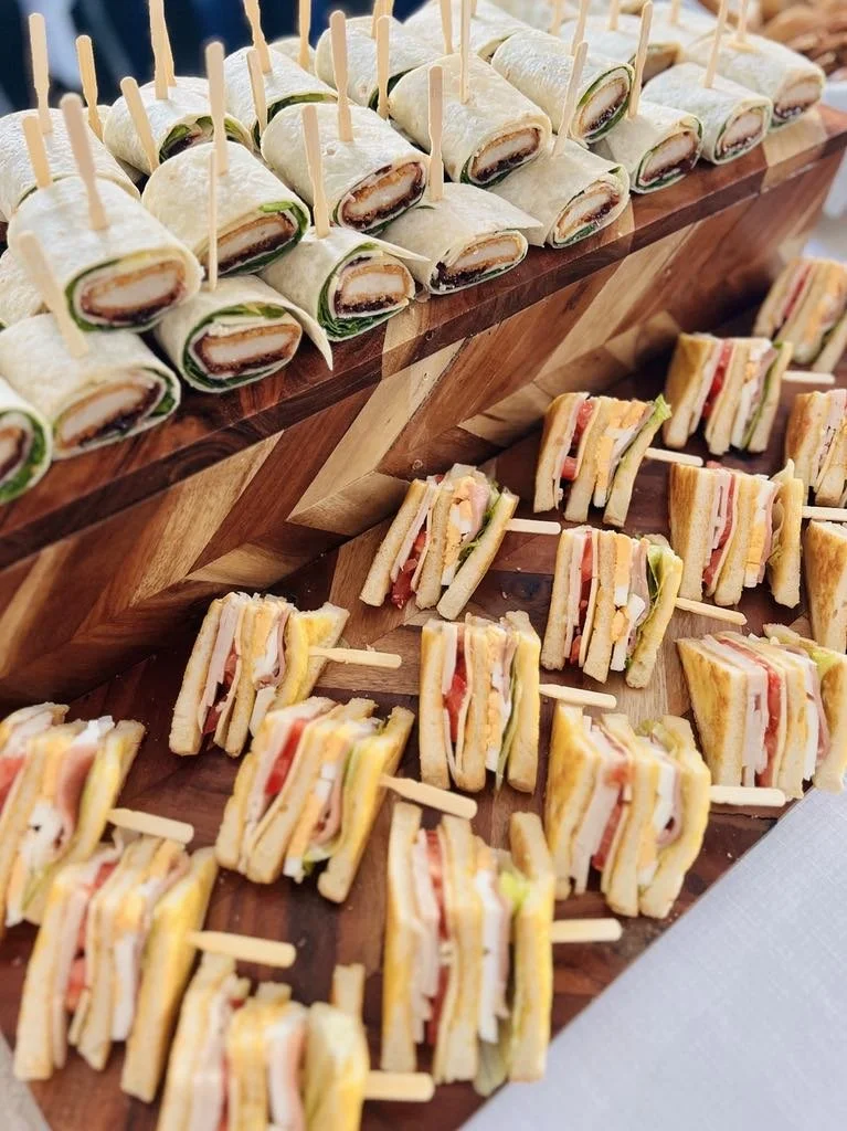Tray of assorted miniature sandwiches, including wrap sandwiches with lettuce and meat and club sandwiches with multiple layers of bread, meat, cheese, and vegetables, all served with toothpicks.