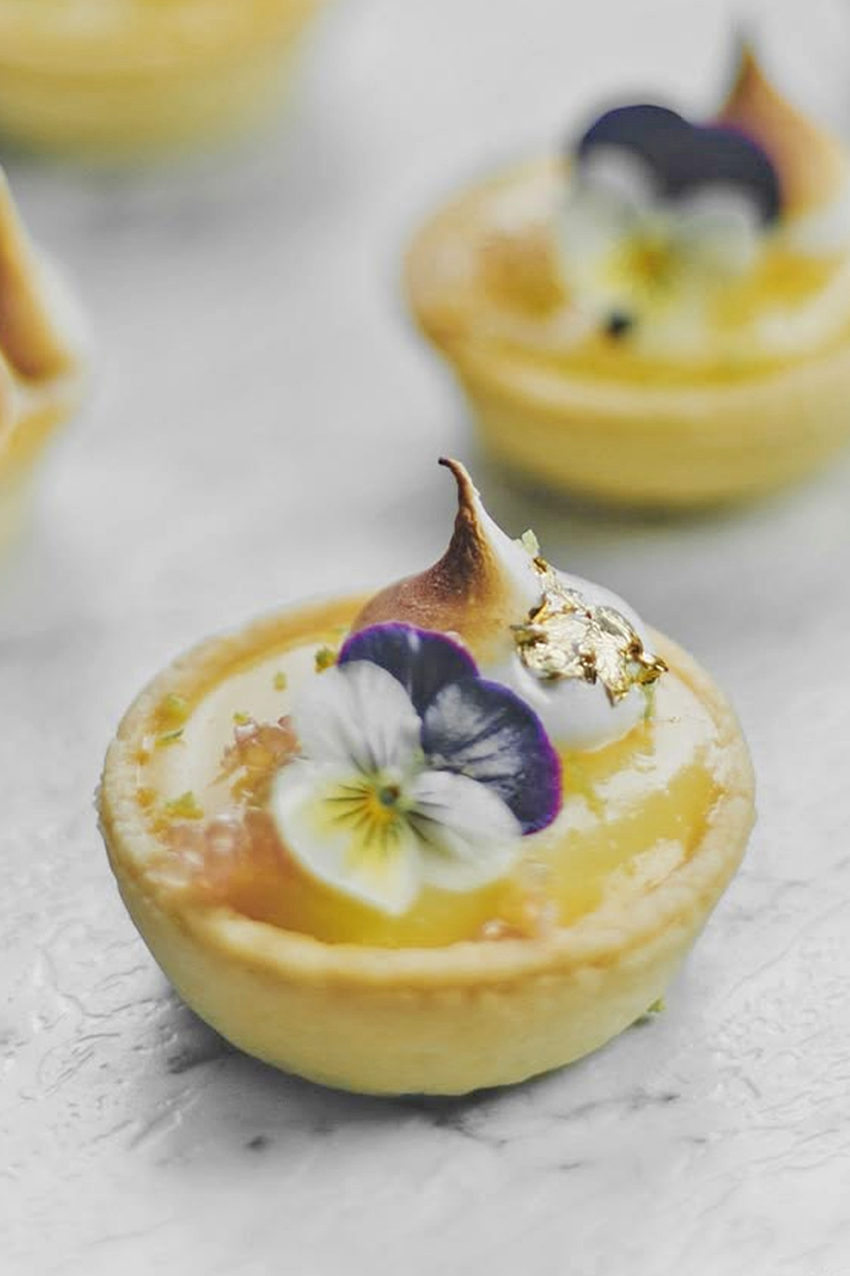 Mini lemon tart topped with edible flowers, gold leaf, and toasted meringue.