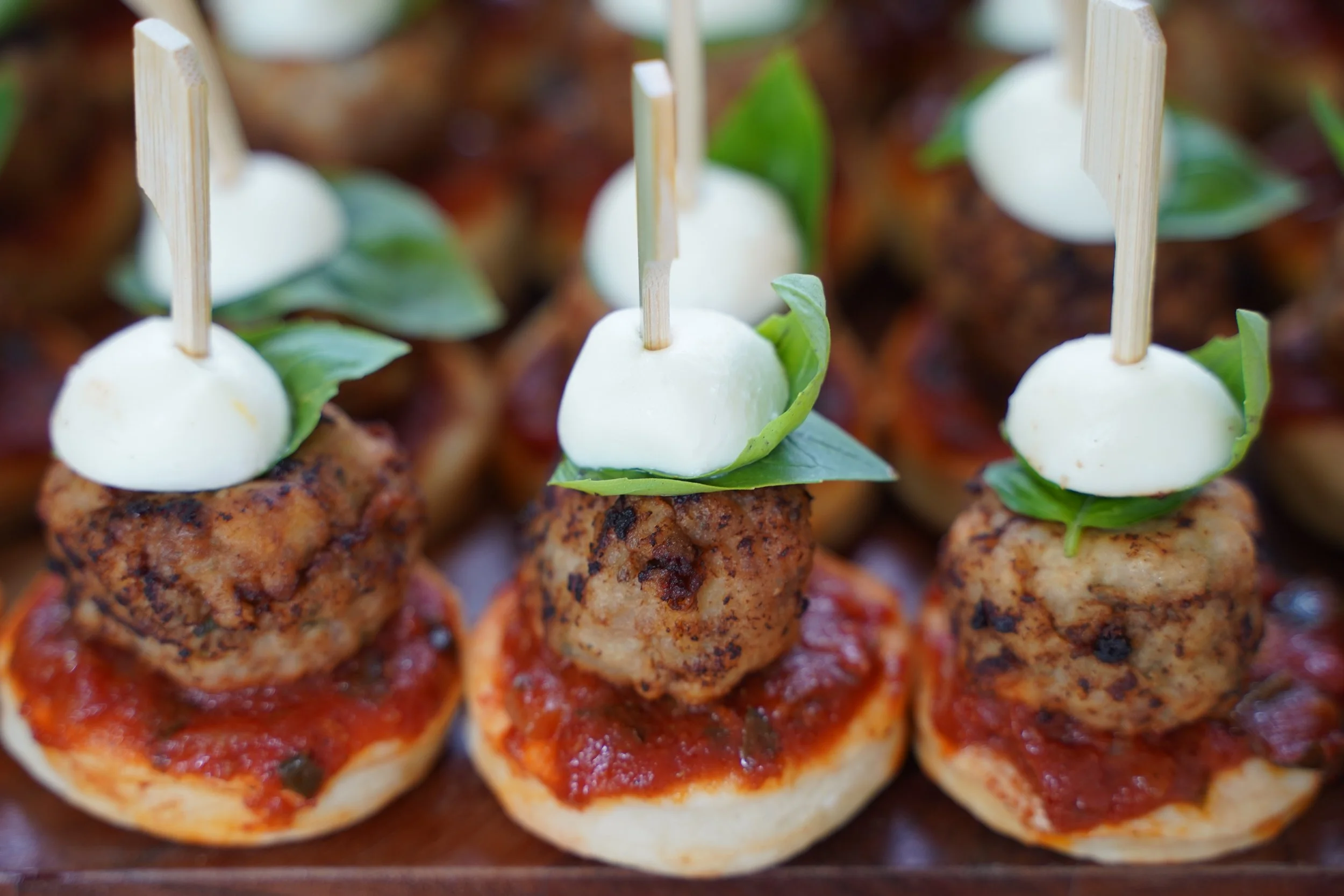 Close-up of mini appetizers with meatballs on tomato sauce, topped with mozzarella cheese, fresh basil, and a bamboo pick.