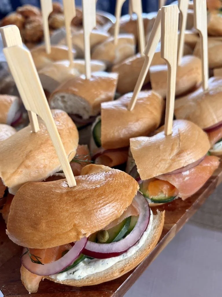 Close-up of small sandwiches with smoked salmon, cucumber, red onion, and cream cheese on mini baguettes, displayed on a wooden tray with wooden toothpicks.