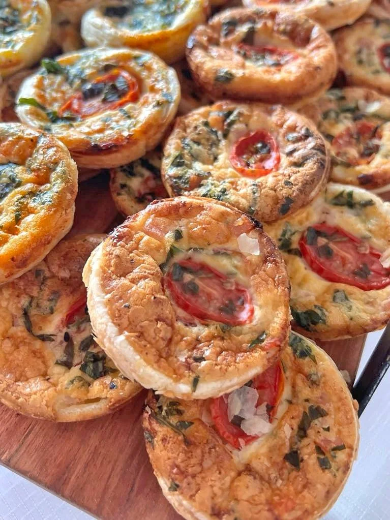 Close-up of miniature savory tarts with tomato, herbs, and melted cheese on a wooden serving board.