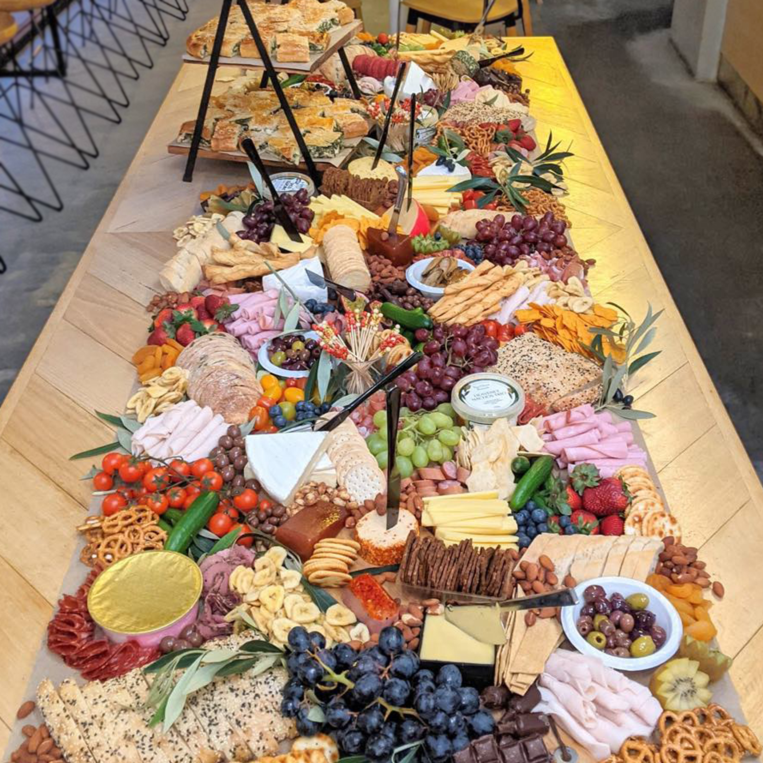 Long wooden table filled with an assortment of cheeses, grapes, crackers, cherry tomatoes, strawberries, and various snacks, with some decorative greenery and serving utensils.