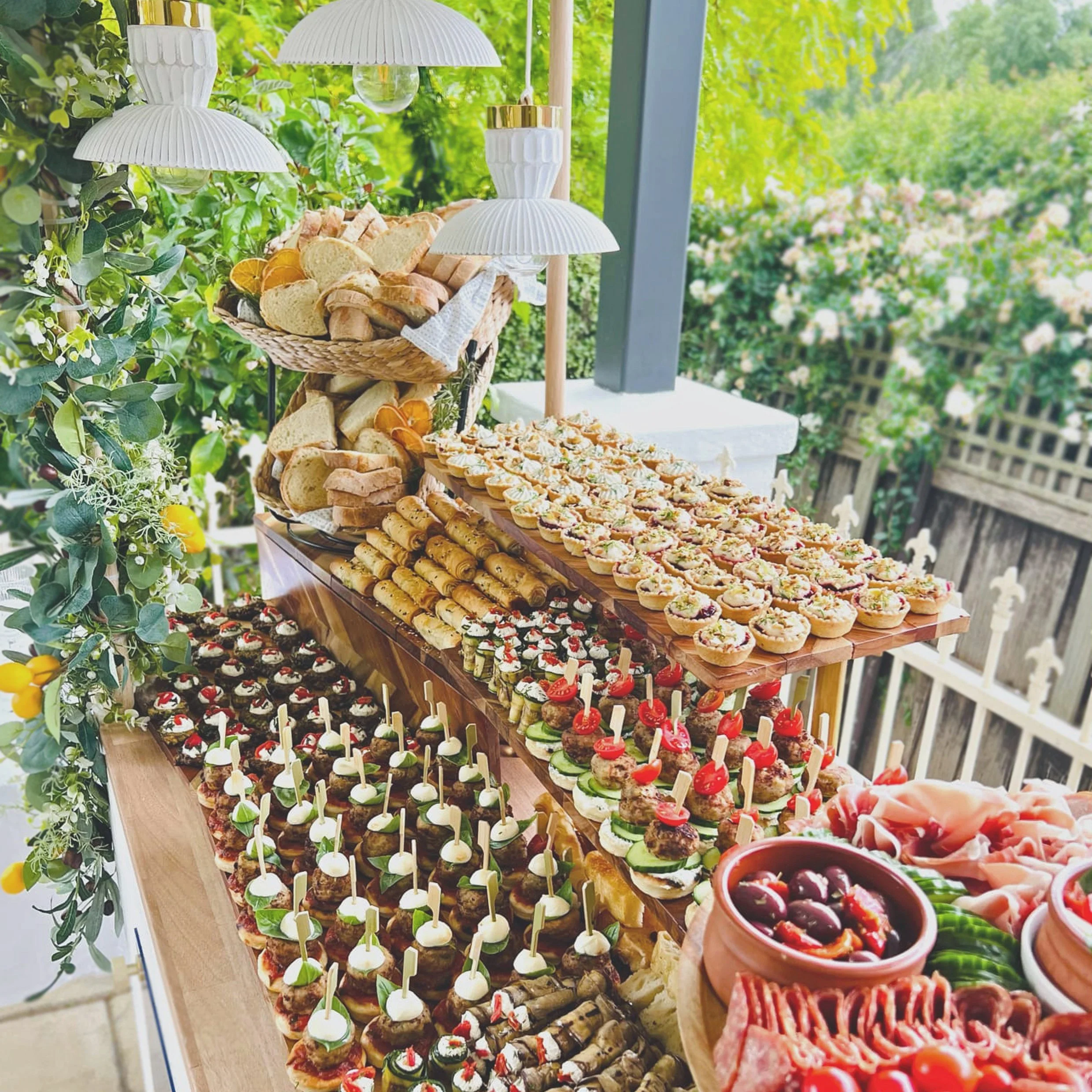 A variety of finger foods and appetizers on a decorated outdoor buffet table, including petite sandwiches, canapés with toppings, skewered snacks, and dips, surrounded by greenery.