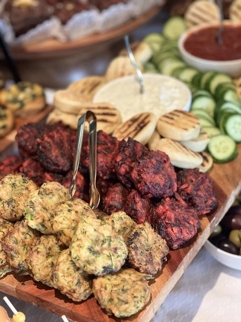 An assortment of grilled meatballs, including yellowish herb meatballs, dark red beef patties, and reddish sausage balls, served with grilled bread slices and fresh cucumber slices on a wooden platter.