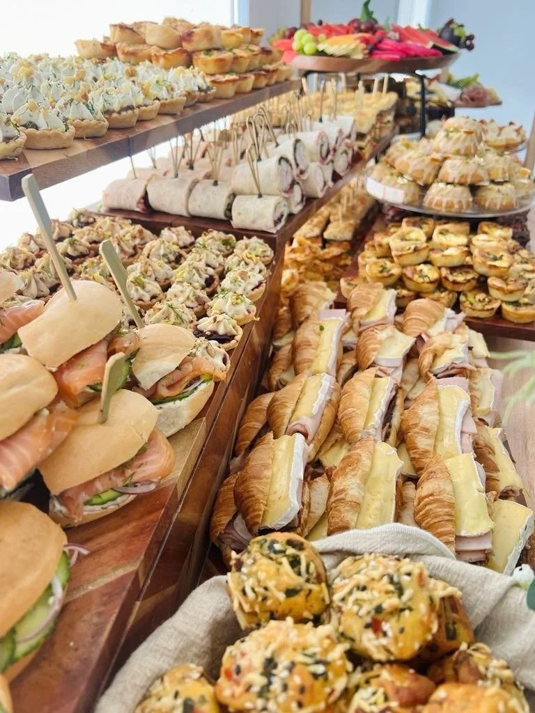 Assorted finger foods including mini sandwiches with ham and cheese, croissant sandwiches, vegetable and meat topped tartlets, fruit and vegetable skewers, and bite-sized pastries on a large table at a buffet or party.