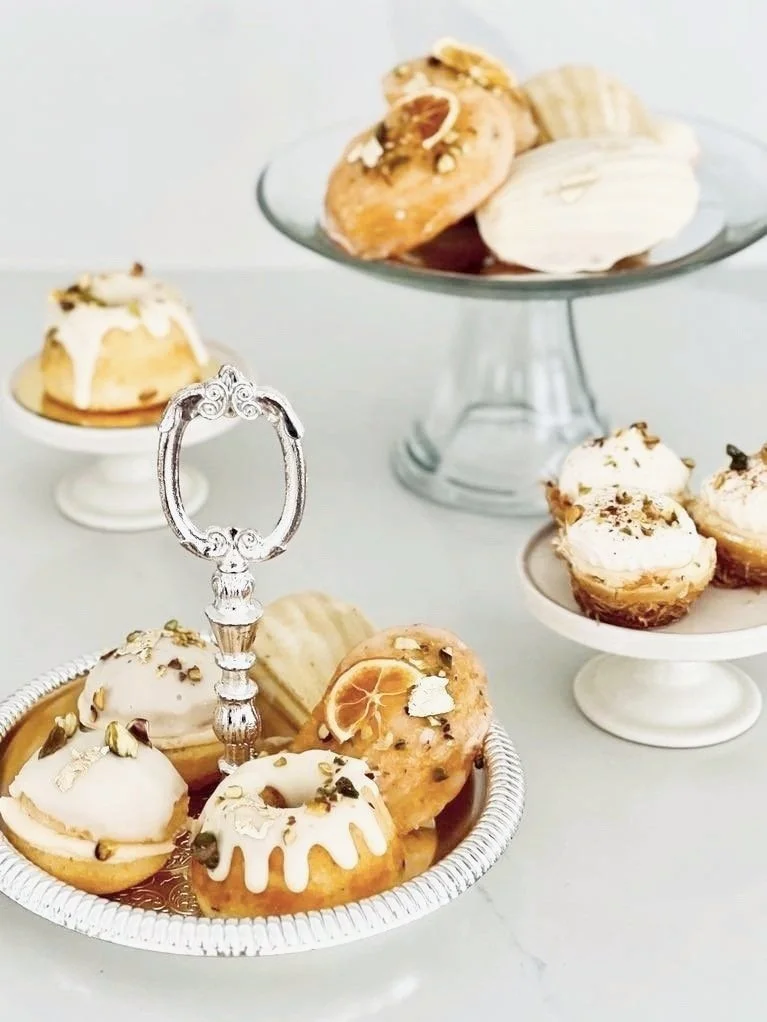 Assorted mini cakes decorated with white icing, nuts, and dried citrus slices, displayed on white and glass cake stands.