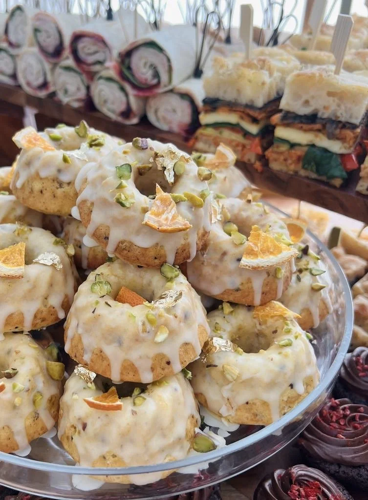 A glass platter filled with glazed mini Bundt cakes topped with chopped pistachios and slivered almonds, with assorted sandwich and dessert items in the background.