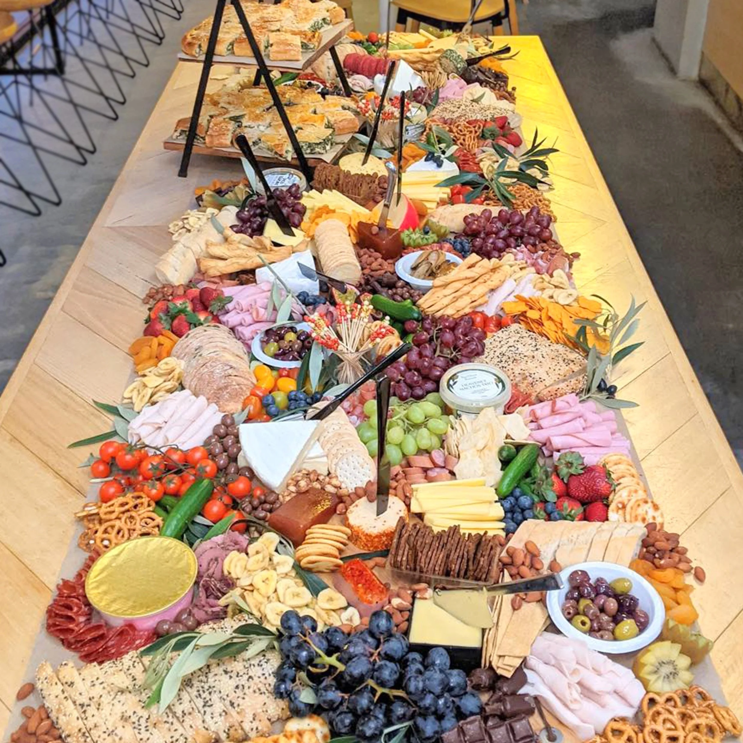 A long wooden table covered with a variety of cheeses, fruits, crackers, vegetables, and meats arranged for a buffet or gathering.