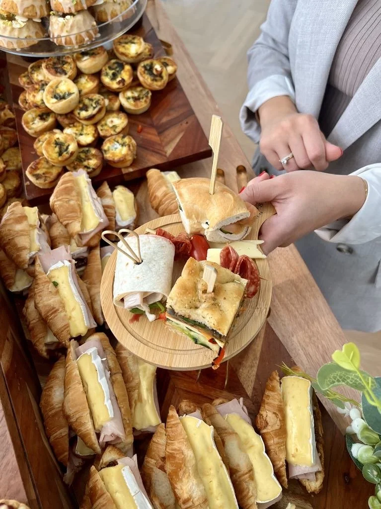 A person holding a small platters of assorted finger foods and snacks, with a display of mini croissant sandwiches, quiches, and small pastries with cheese in the background.