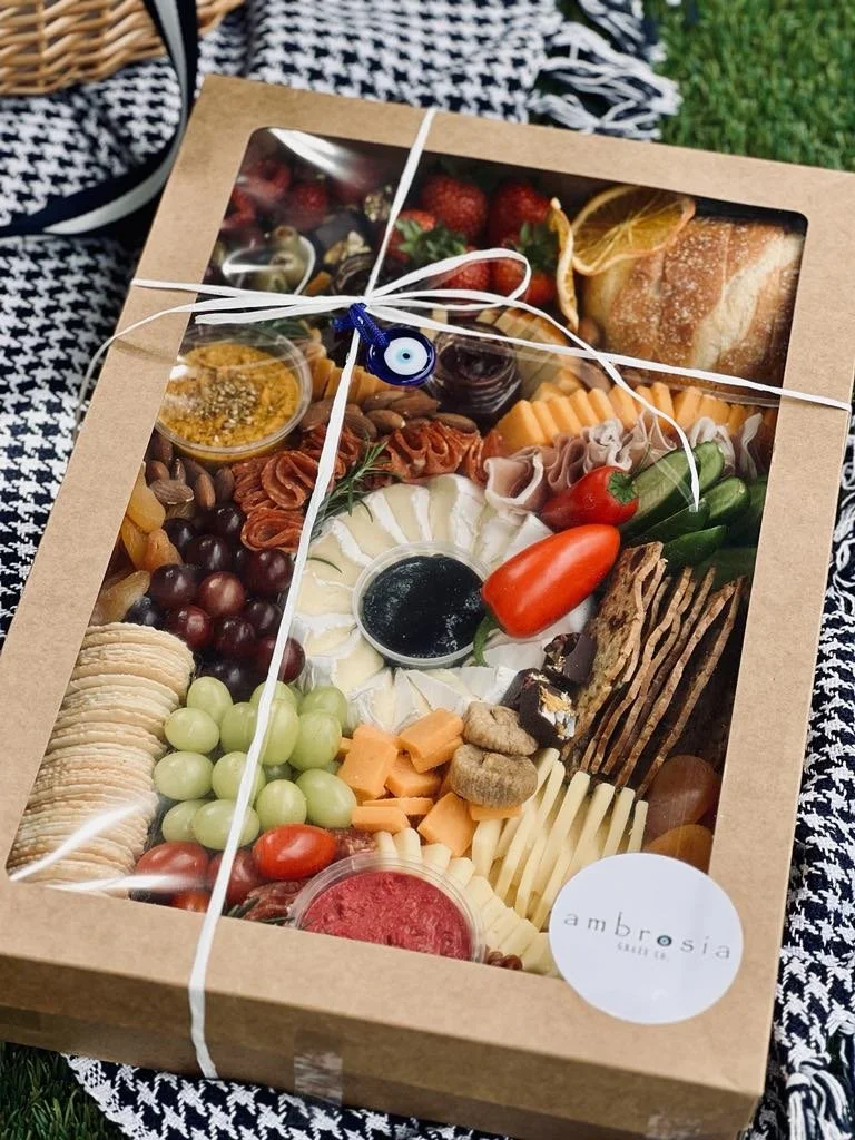 Gift box of assorted cheeses, grapes, cherry tomatoes, cured meats, crackers, strawberries, and dips, wrapped with white and blue twine and placed on a checkered cloth outdoors.