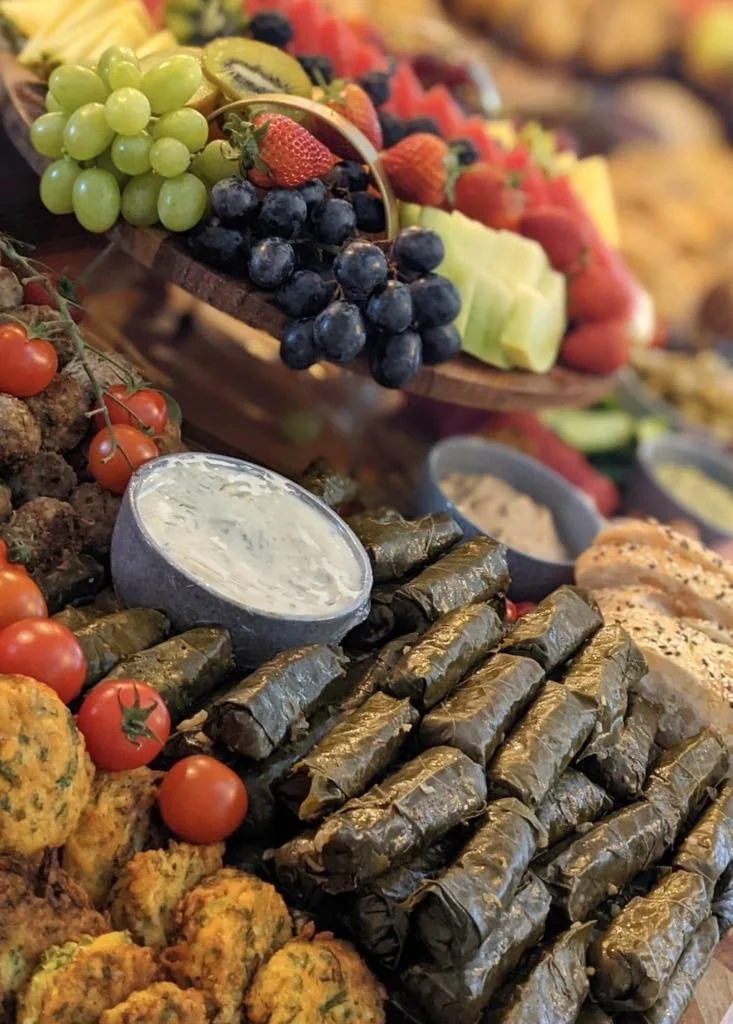 Assorted fresh fruits, grape leaves rolled in vine, cherry tomatoes, vegetable falafel, and creamy dipping sauce on a buffet table.