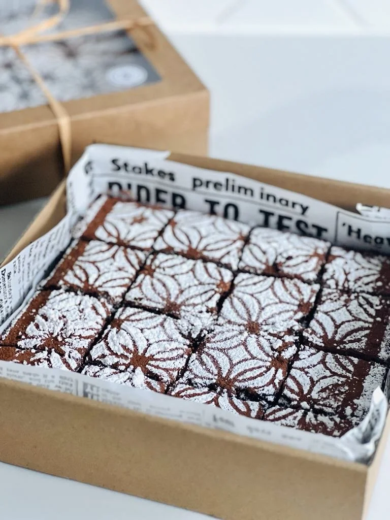 A box of decorated chocolate brownies topped with powdered sugar, arranged in a grid pattern, lined with newspaper