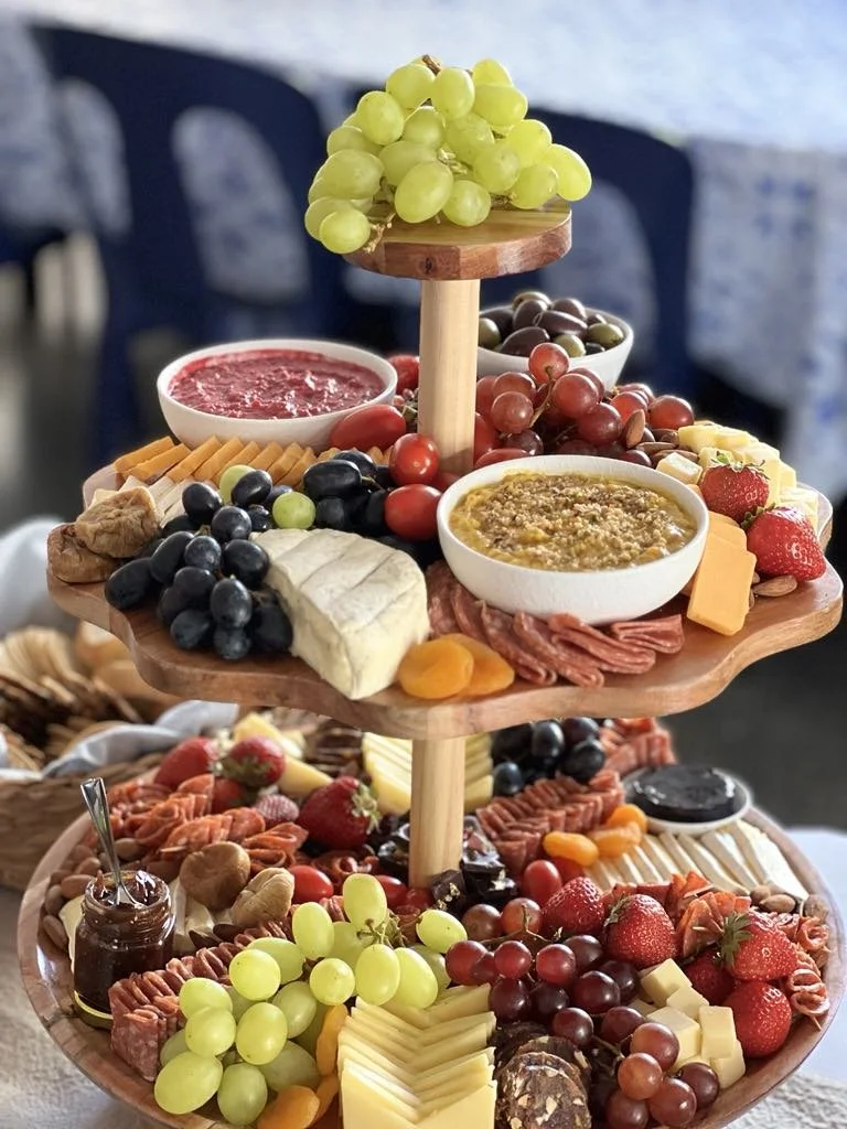 A three-tiered wooden serving tray filled with grapes, cheese, strawberries, and various charcuterie, set on a table with a blurred background of chairs and a tablecloth.