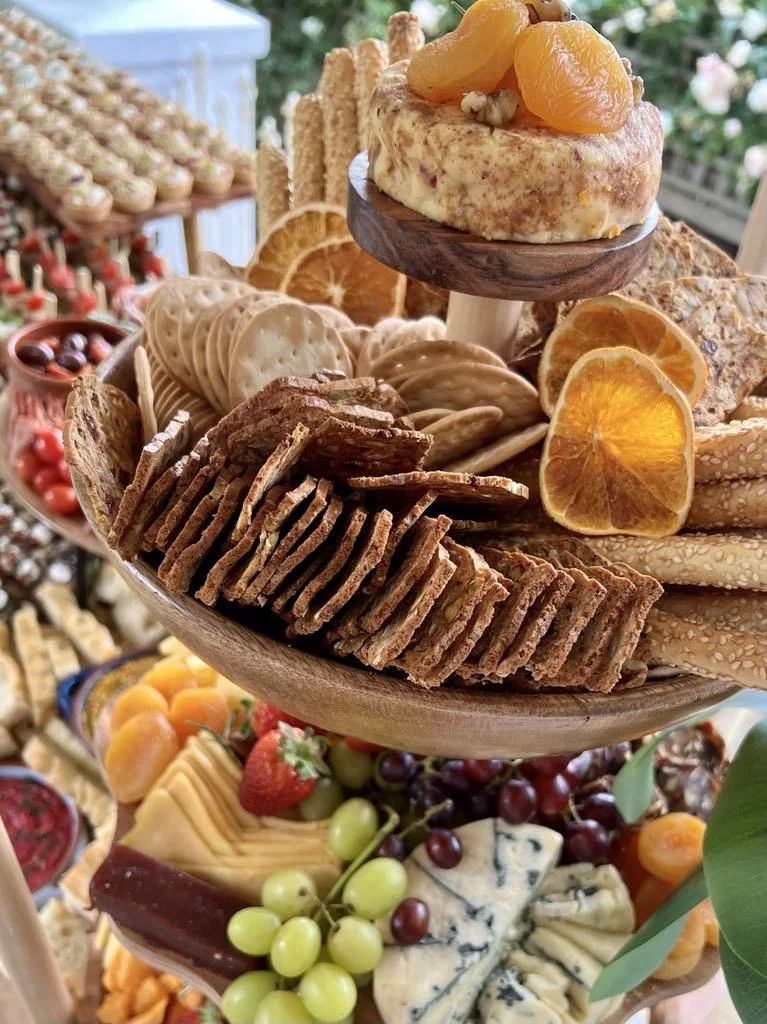 A tiered platter of assorted desserts and snacks, including cookies, crackers, dried fruit, cheese, fruit, and a small cheesecake topped with fruit.