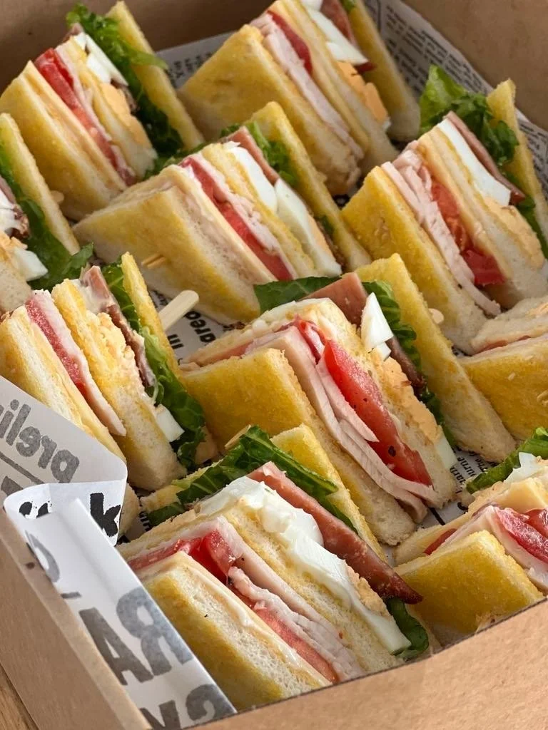 Tray of assorted club sandwiches with lettuce, tomato, turkey, ham, cheese, and white bread, arranged in a cardboard box lined with newspaper-style paper.