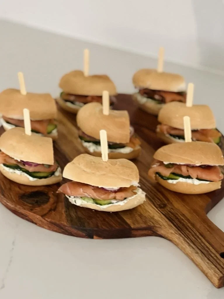 Mini sandwiches with smoked salmon, cream cheese, and cucumber on mini sandwich buns, skewered with toothpicks, arranged on a wooden serving board.