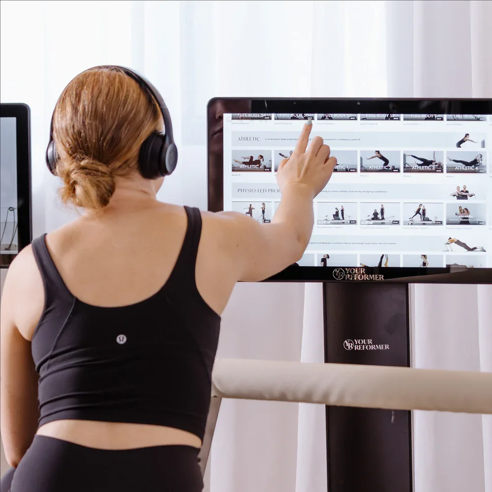 Woman wearing headphones on a pilates reformer selecting pilates workout images on a computer screen
