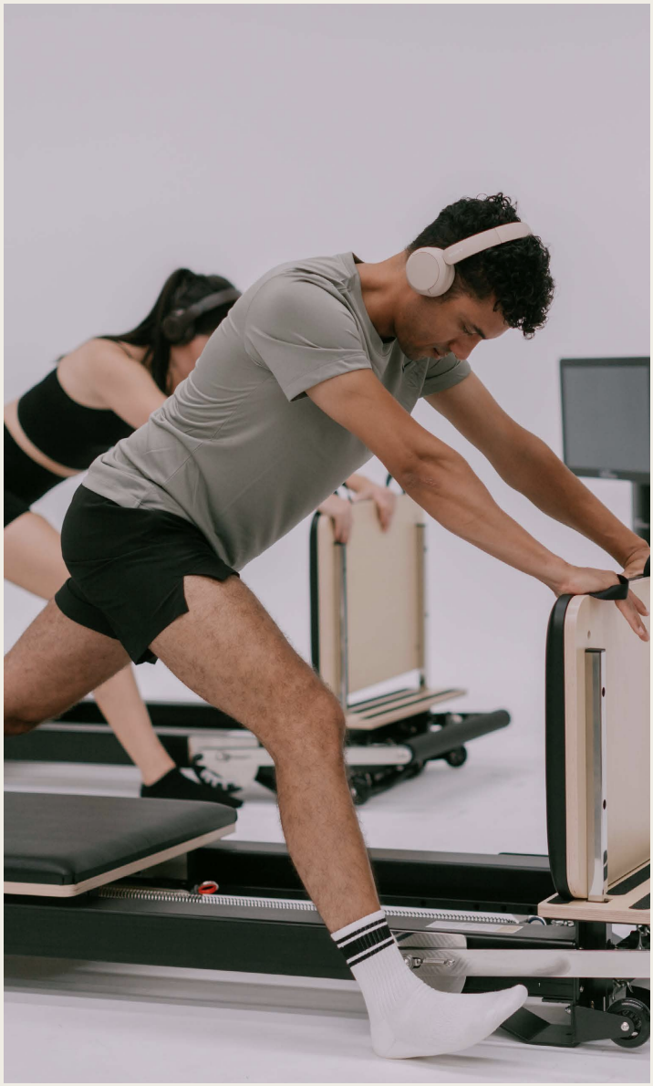 Man wearing headphones stretching on a Pilates reformer machine during workout, with woman in the background also exercising.