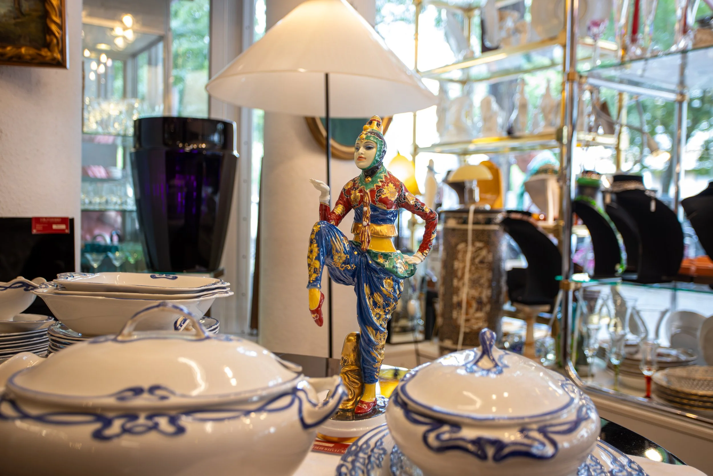 Deko-Figur eines tanzenden asiatisch wirkenden Mannes, umgeben von Porzellanschüsseln, in einem Laden mit Regalen und Geschirr im Hintergrund.