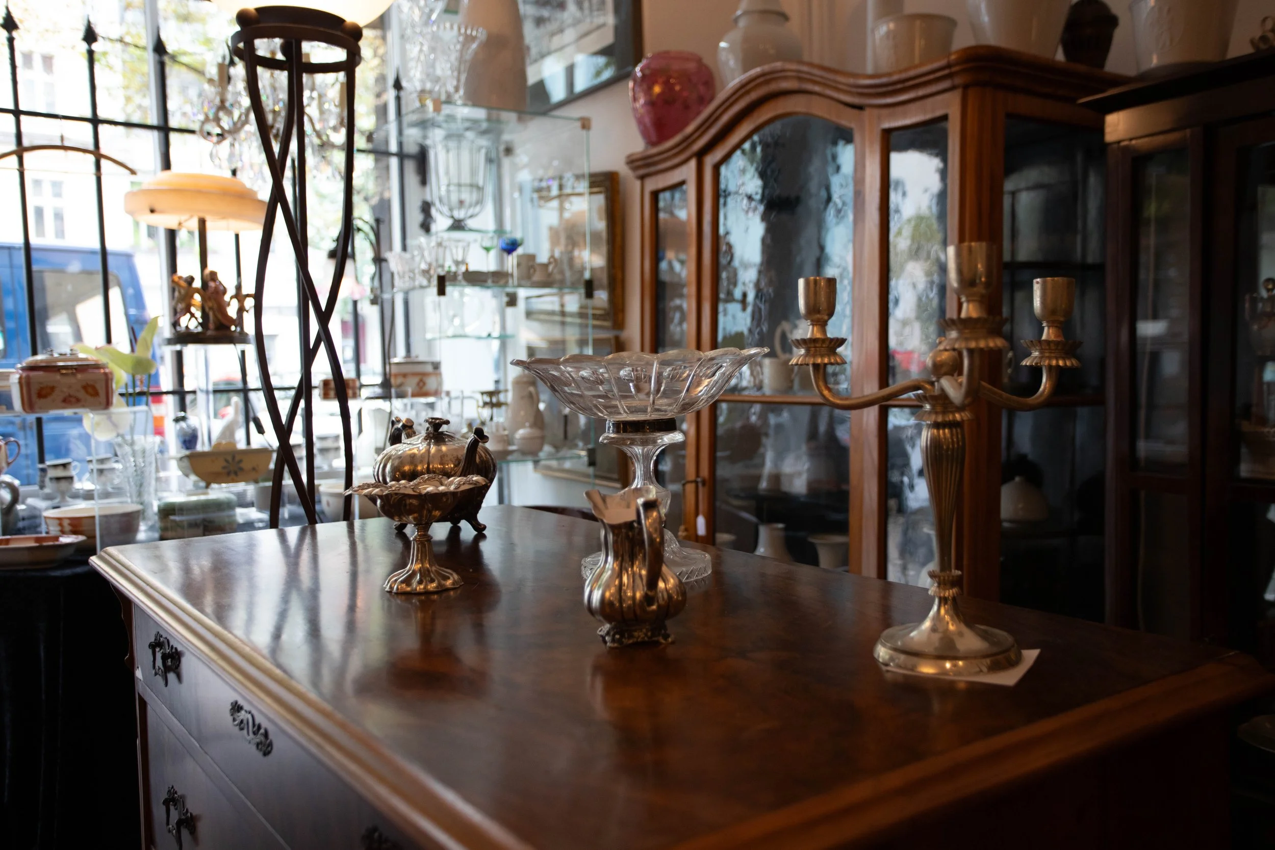 Innenansicht eines Antiquitätengeschäfts mit Holzmöbeln und Schmuckstücken, darunter Kerzenhalter, Vasen und Glasdekorationen.