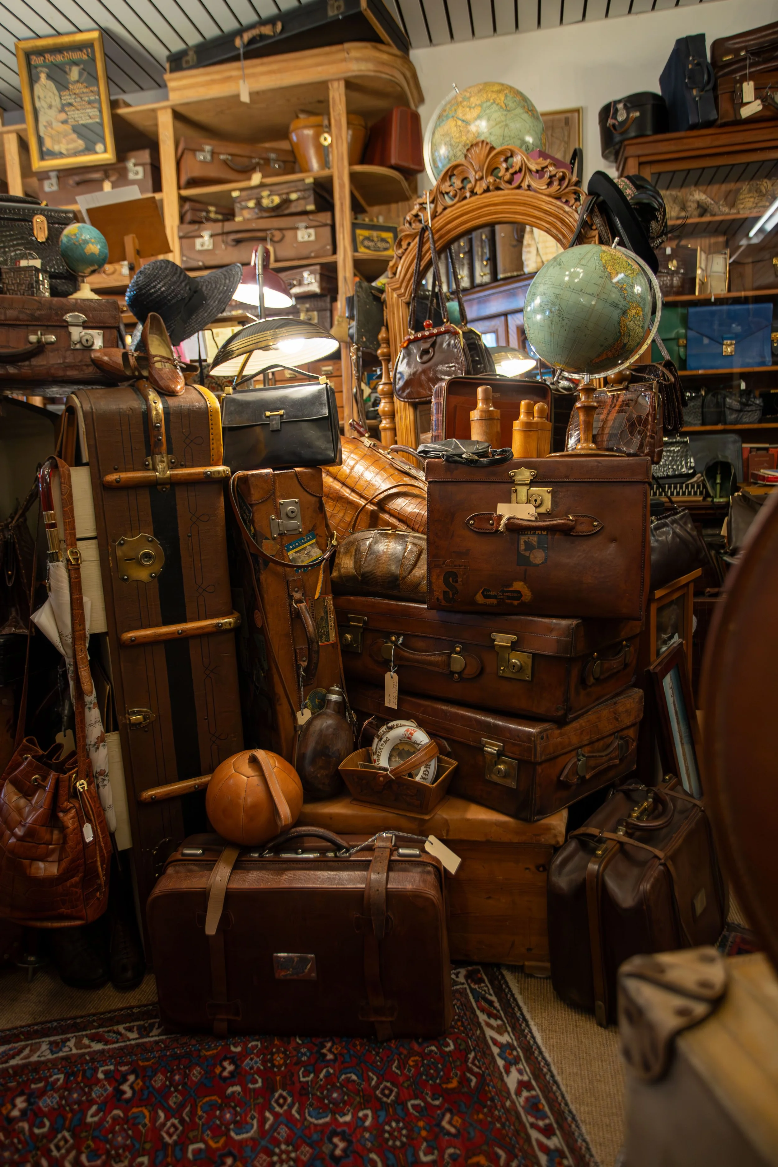 Haufen alter Lederkoffer, Taschen und Businesstaschen in einem Antiquitätengeschäft, mit Globus, Vintage-Spiegel und anderen Dekorationsgegenständen im Hintergrund.