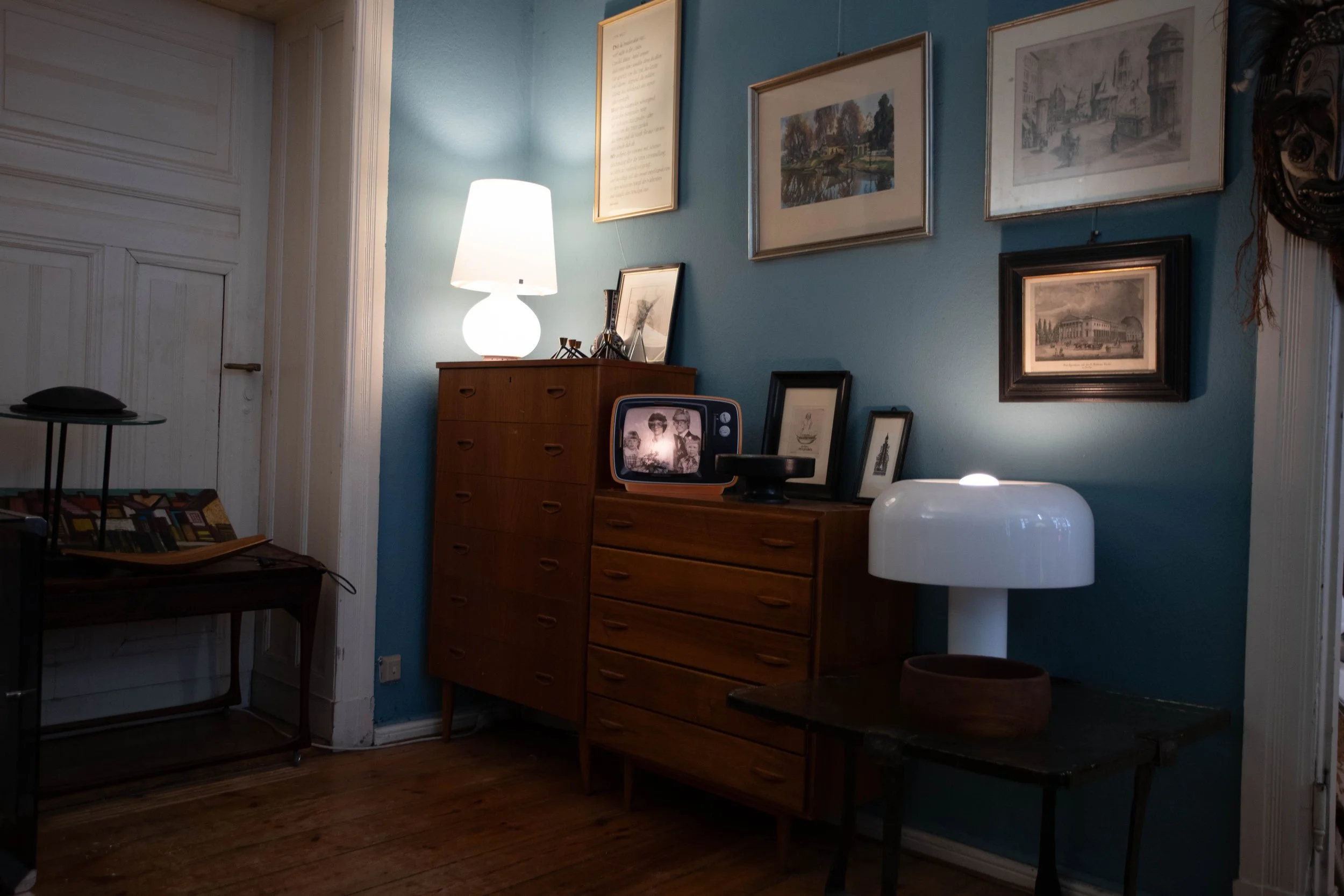 Ein Wohnzimmer mit Holzfußboden, blauer Wand und verschiedener Kunst an der Wand, eine weiße Tischlampe, ein alter Fernseher auf einem Holzschrank und weitere Dekorationsobjekte.