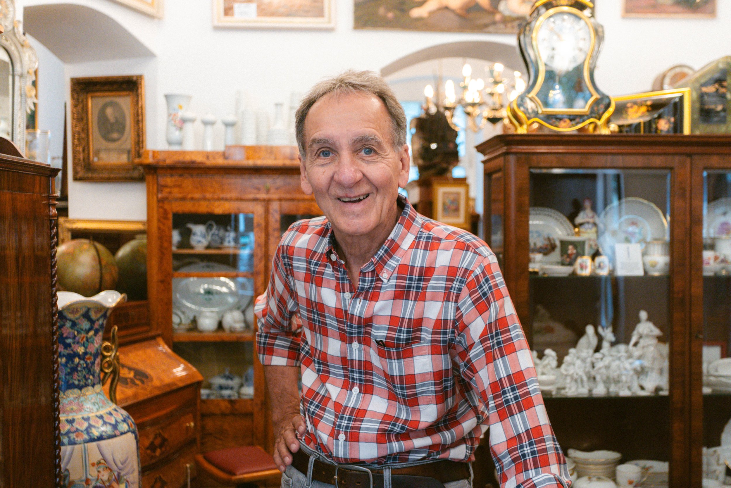 Ein älterer Mann trägt ein rotes kariertes Hemd und steht in einem Antiquitätengeschäft mit Holzvitrinen voller alten Porzellans und Dekorationsgegenständen.