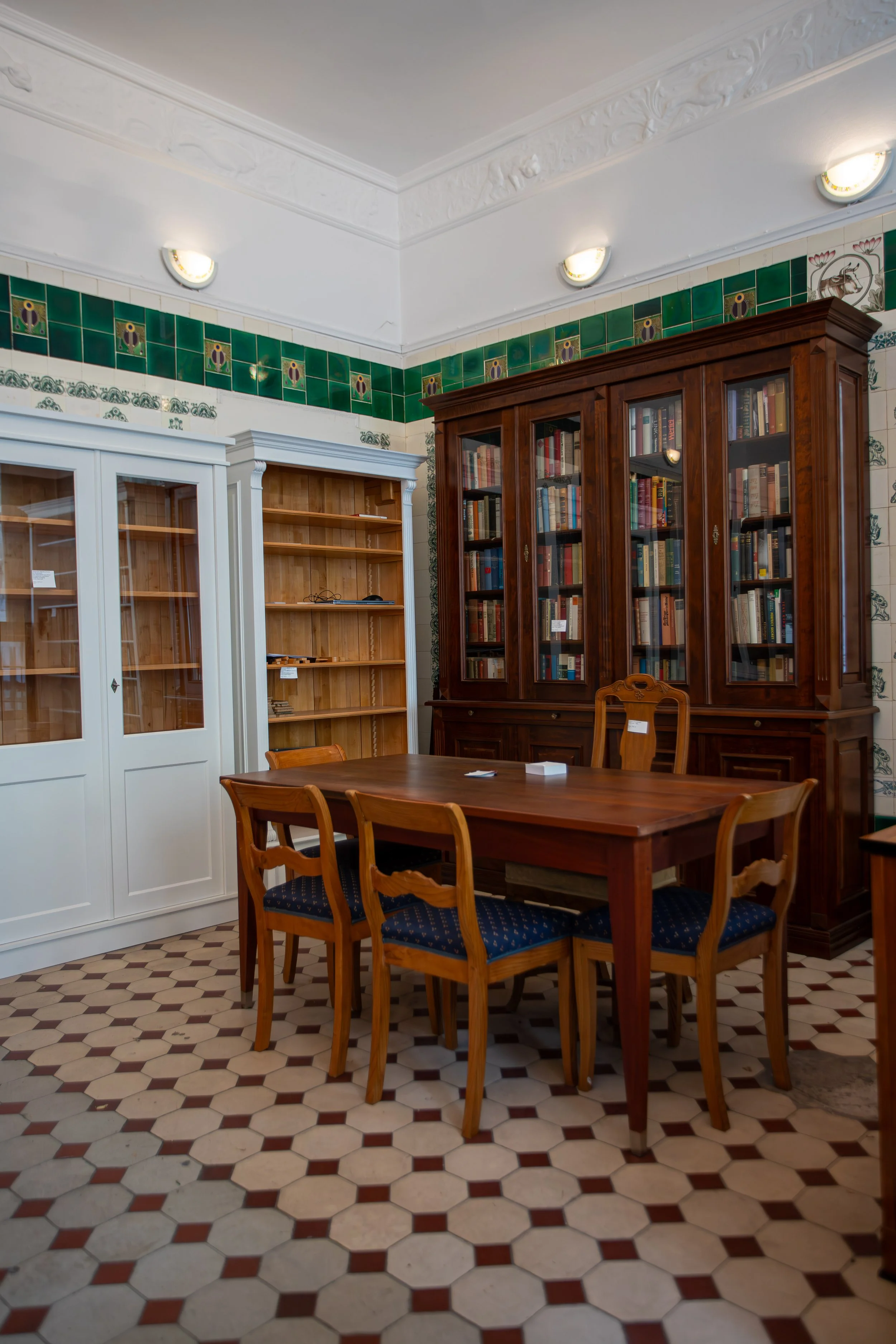 Ein Raum mit einem Holztisch, sechs Holzstühlen, einer großen Holzbibliothek mit Glasschrank, einigen leeren Bücherregalen und bemalten Fliesen an der Wand.
