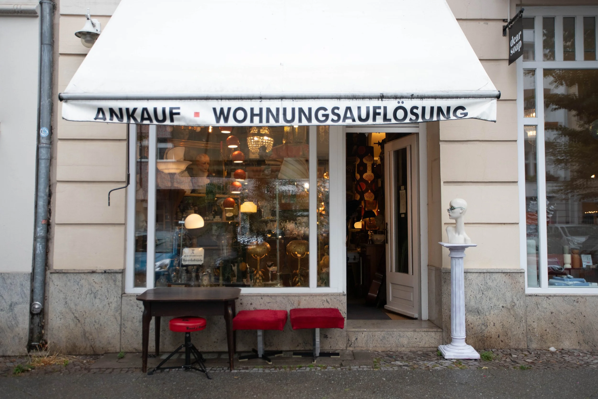 Verkaufsraum eines Antiquitätenladens mit Lampen, einem Dummkopf-Statue und Möbeln. Außenansicht mit Beschriftung des Geschäfts: 'ANKAUF · WOHNUNGSaufsLÖSUNG'.