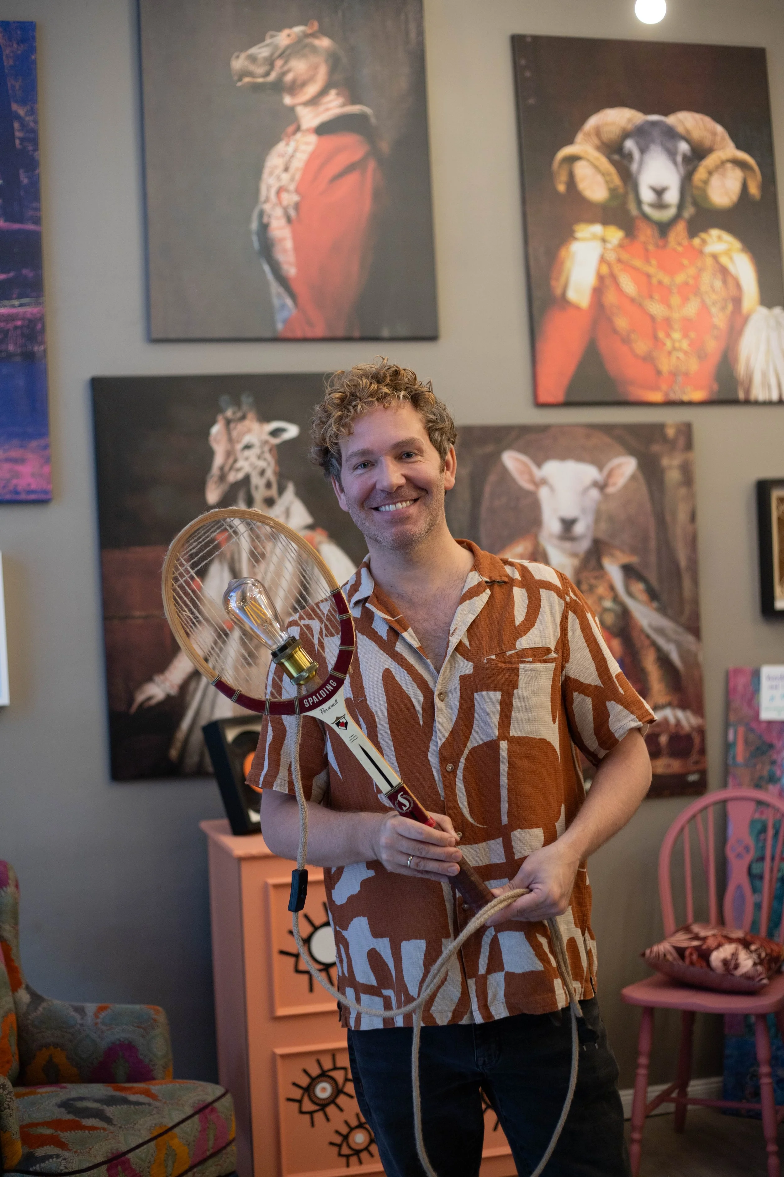 Ein lachender Mann in einem braun-weißen Hemd hält einen Tennisracket mit einer Glühbirne und einem Kabel darin. Im Hintergrund sind bunte Tierporträts an der Wand und ein pinker Stuhl.