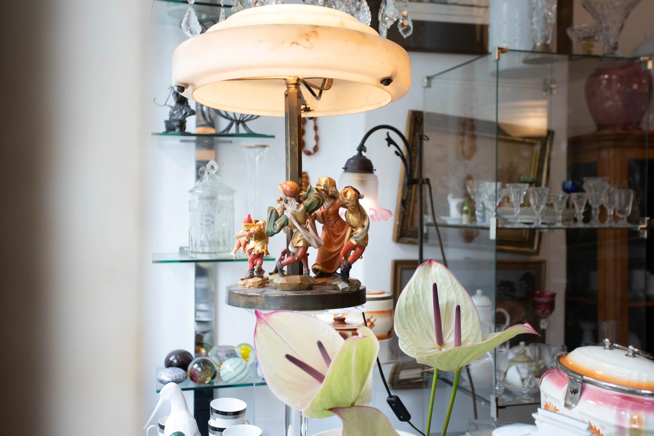 Ein Vintage-Tischlampenschirm mit einer Figurengruppe darunter, auf einem Glasregal mit Kristallvasen und anderen Dekorationsgegenständen, sowie zwei große weiße Blumen im Vordergrund.