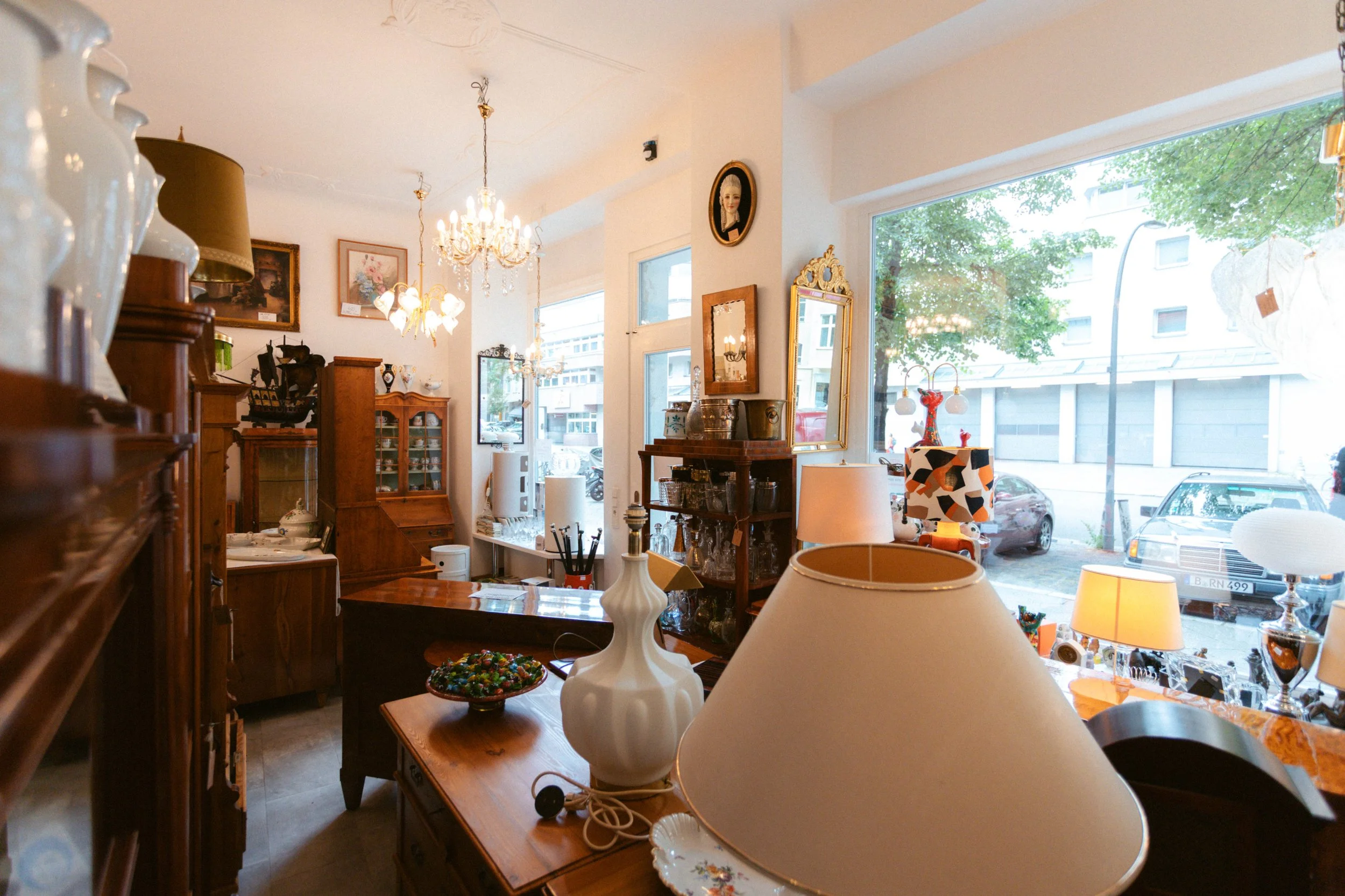 Ein gemütlicher Innenraum eines Antiquitätengeschäfts mit alten Möbeln, Lampen und Dekorationsgegenständen, durch große Fenster blickt man auf eine Straße mit parkenden Autos.