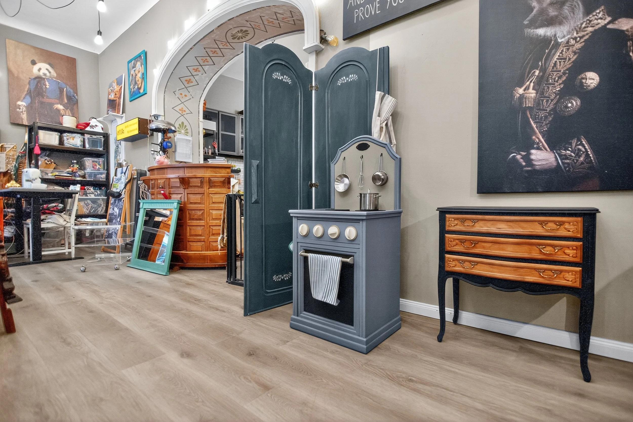 Innenraum mit Spielküche, antiker Kommode, Wandbildern und Möbeln in einem surrealistisch dekorierten Raum.