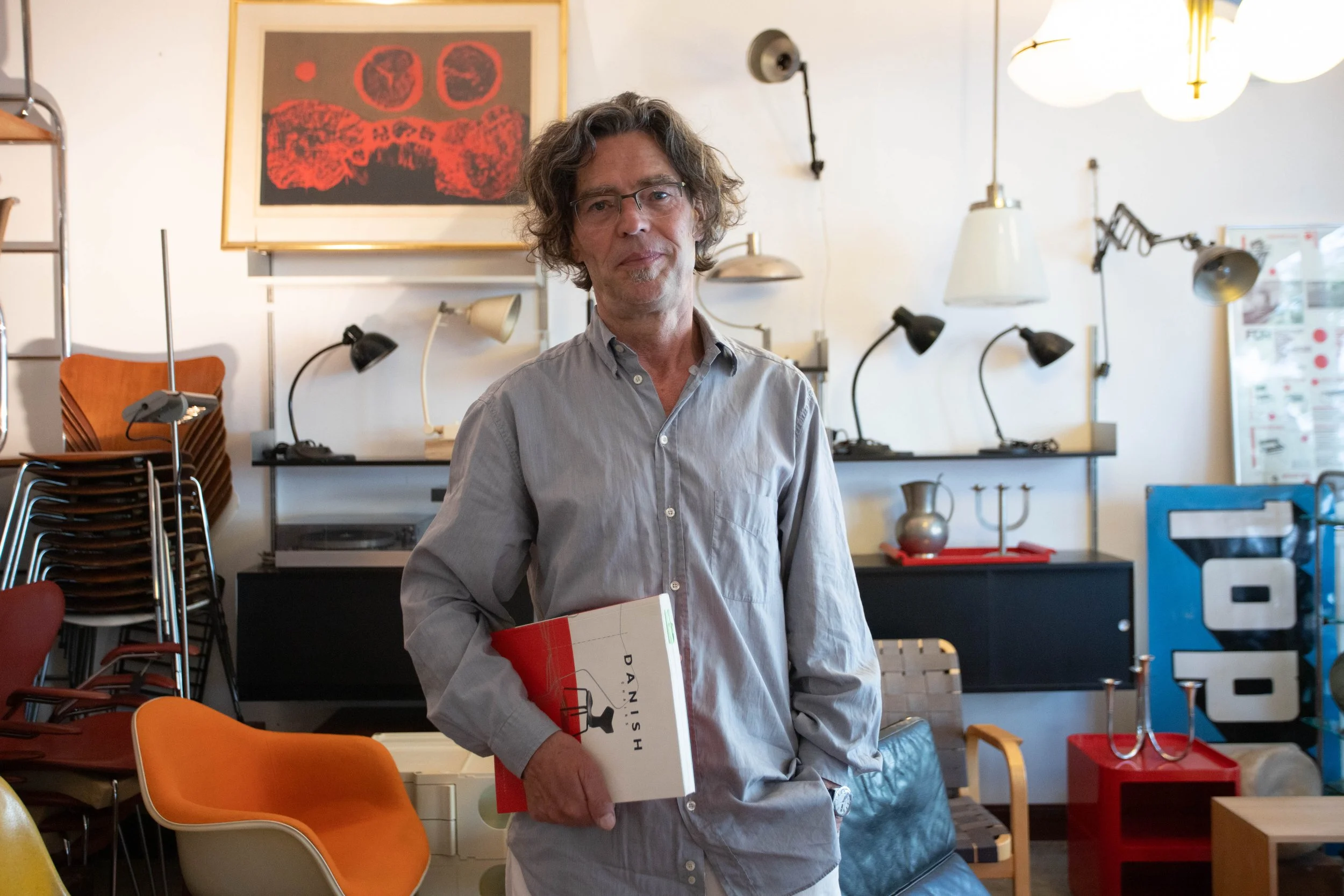 Ein Mann mit Brille, grauem Hemd, steht in einem Raum mit vielen Lampen und Stühlen, hält ein Buch mit dem Titel 'Danish'.