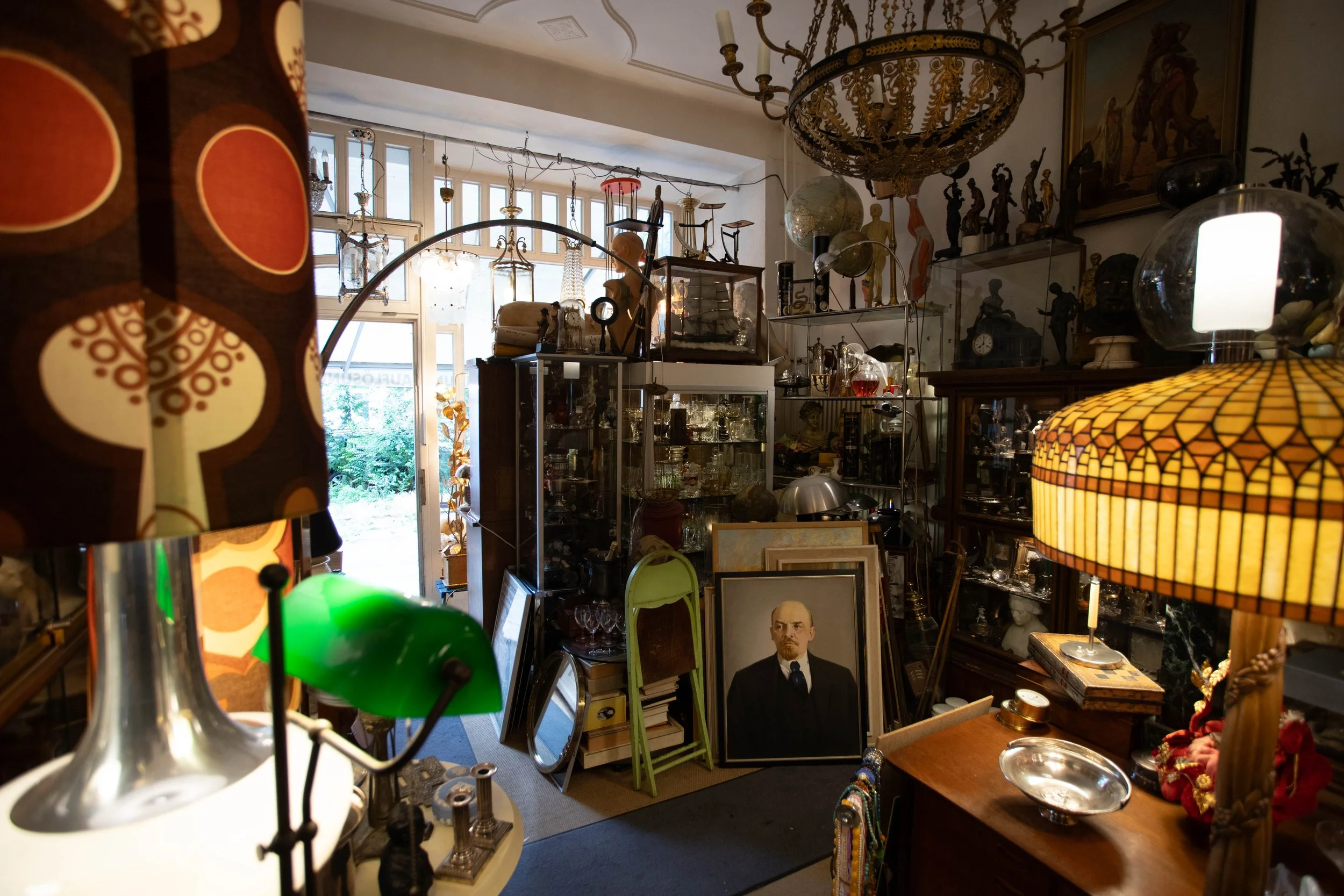 Ein chaotischer Antiquitätenladen voller alter Gegenstände, Gemälde, Lampen und Skulpturen, mit einer offenen Tür im Hintergrund, die auf einen Garten hinausführt.