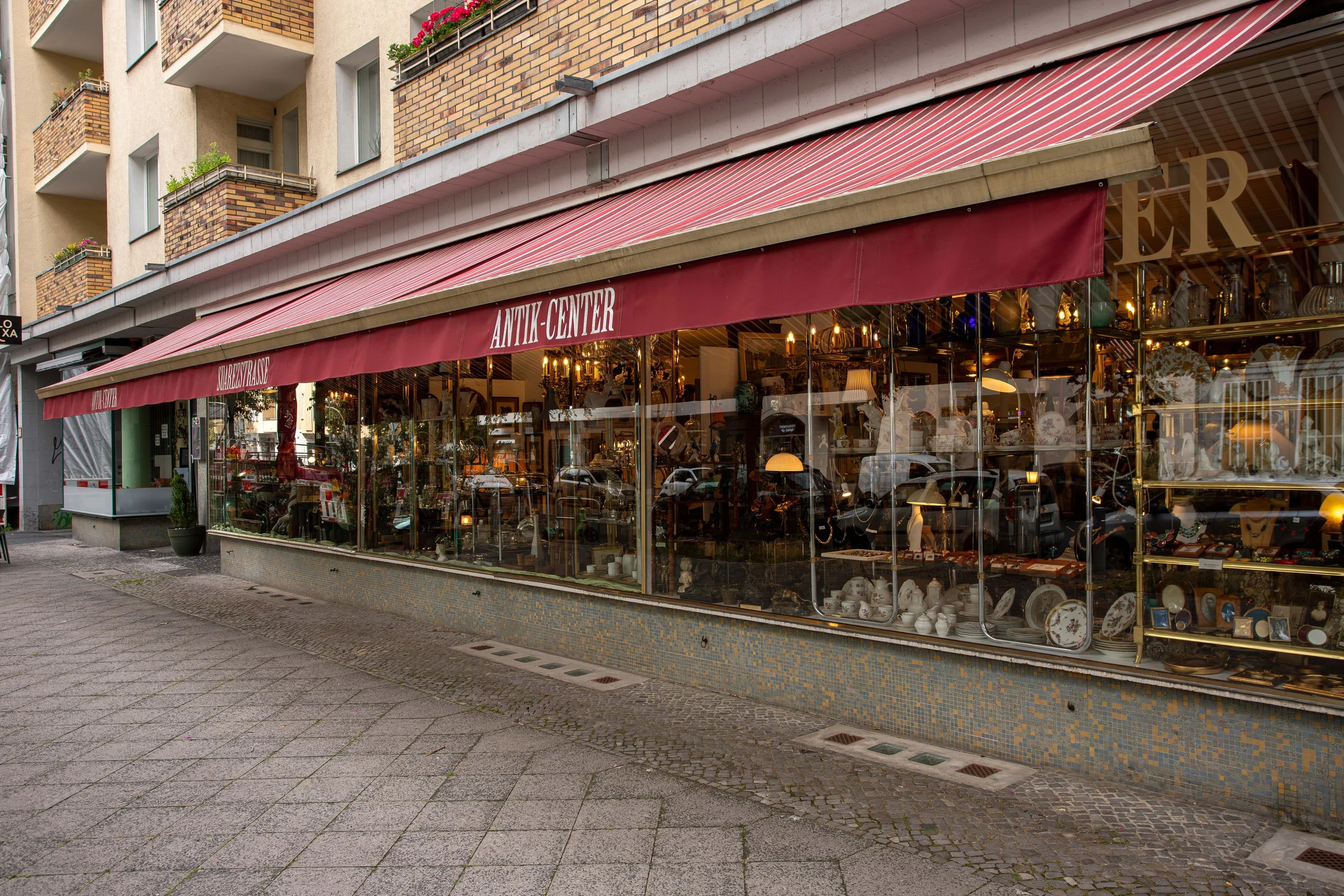 Schaufenster eines Antikladens mit alten Geschirr und Dekorationsartikeln, rotes Markisentuch mit weißen schriftzug 'ANTIK-CENTER' und 'HERITAGE'.