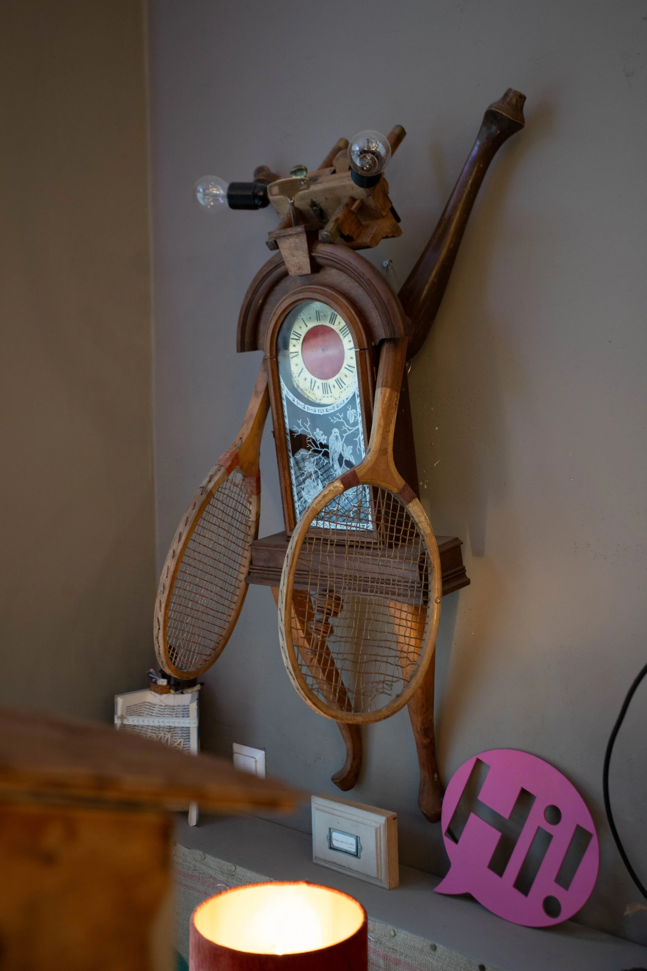 Wanddekoration mit einer Kombination aus alten Tennisschlägern, einem Uhrgehäuse, einem Spiegel mit einem Vogel- und Baumdesign, und einer Lampe mit einem pinkfarbenen Schriftzug 'Hi!' auf dem Boden lehnend gegen die Wand.