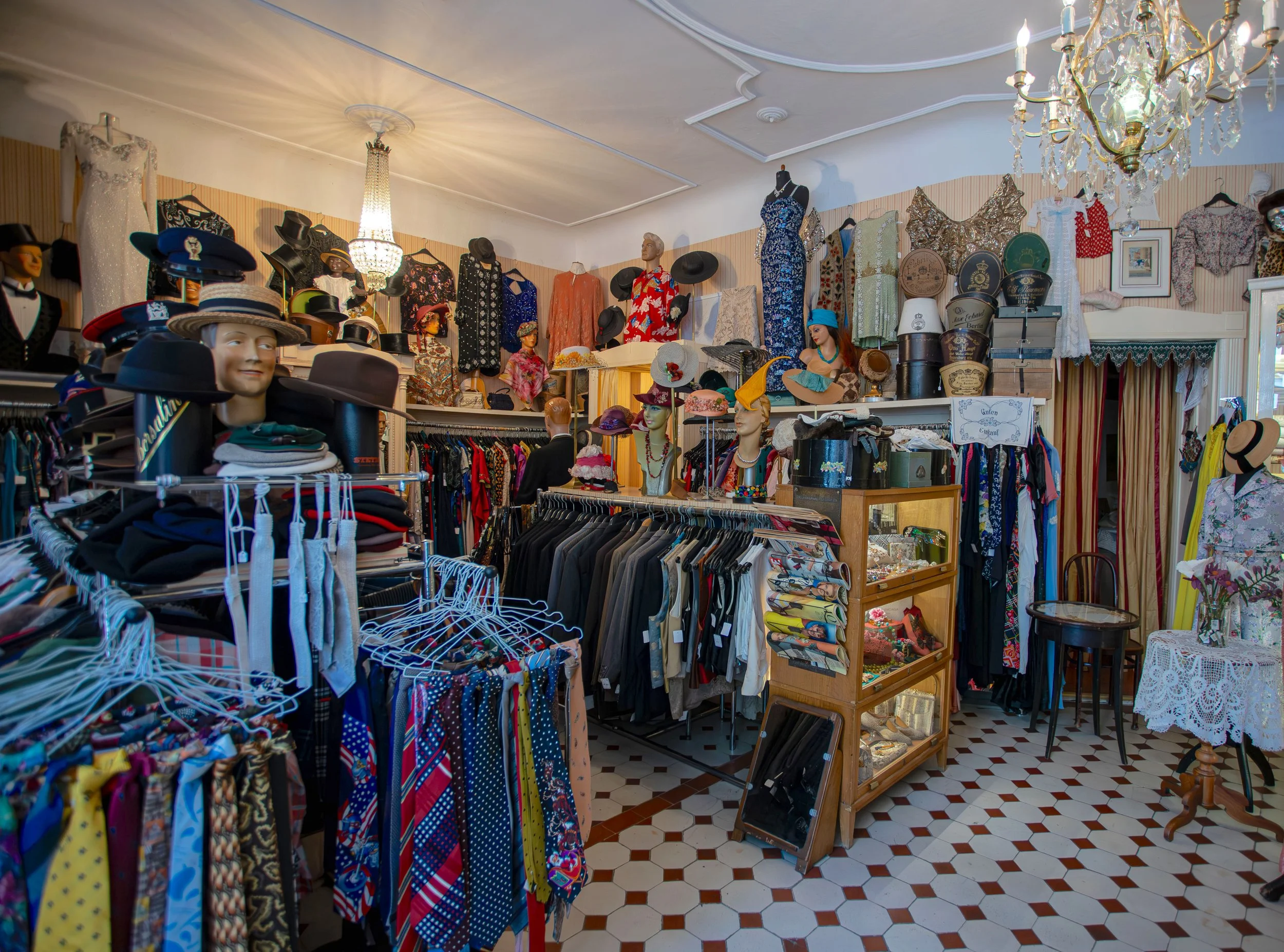 Ein Secondhand-Laden mit Kleidung, Hüten und Accessoires auf Regalen und Kleiderständern, dekoriert mit Kronleuchtern an der Decke.
