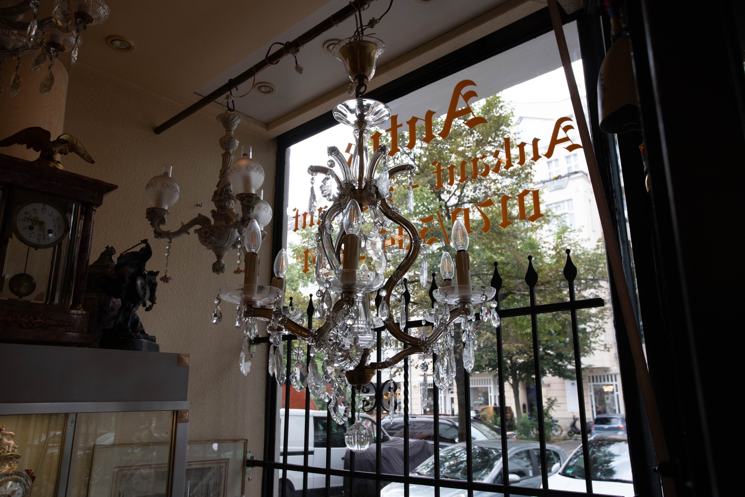 Ein Kronleuchter mit Kristallen hängt vor einem Fenster, auf dem goldfarbener Schriftzug zu sehen ist. Im Hintergrund sind Autos und Bäume sichtbar.