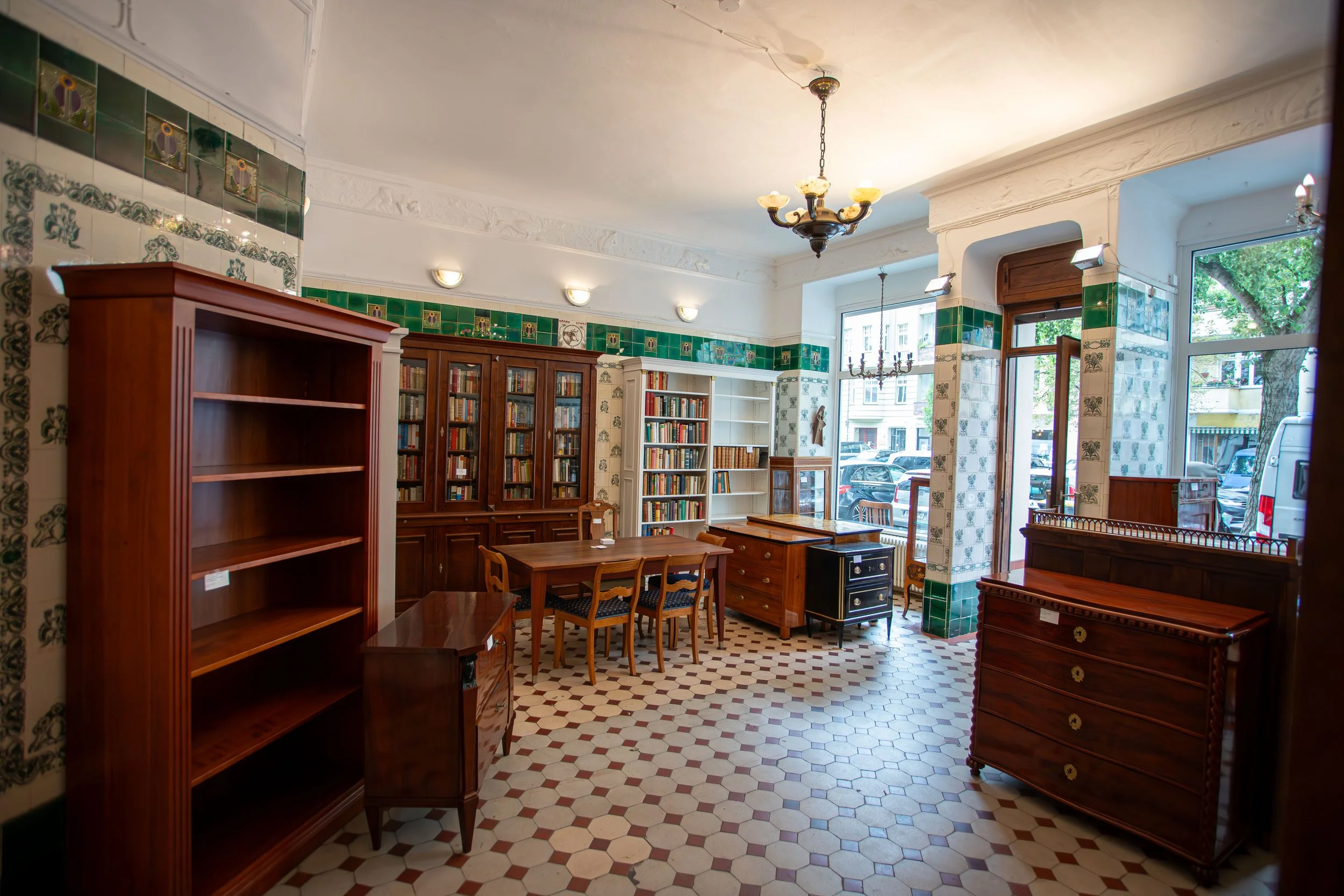 Innenansicht einer Bibliothek mit alten Möbeln, Bücherregalen und großen Fenstern, Tageslicht und grün-weißen Kachelwänden.