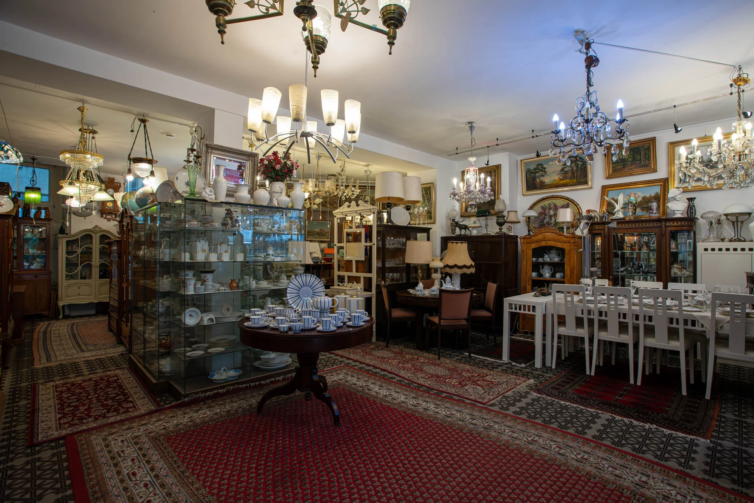 Ein Raum voller antiker Möbel, Porzellan und Gemälde, beleuchtet durch Kristalllampen und Deckenleuchten.