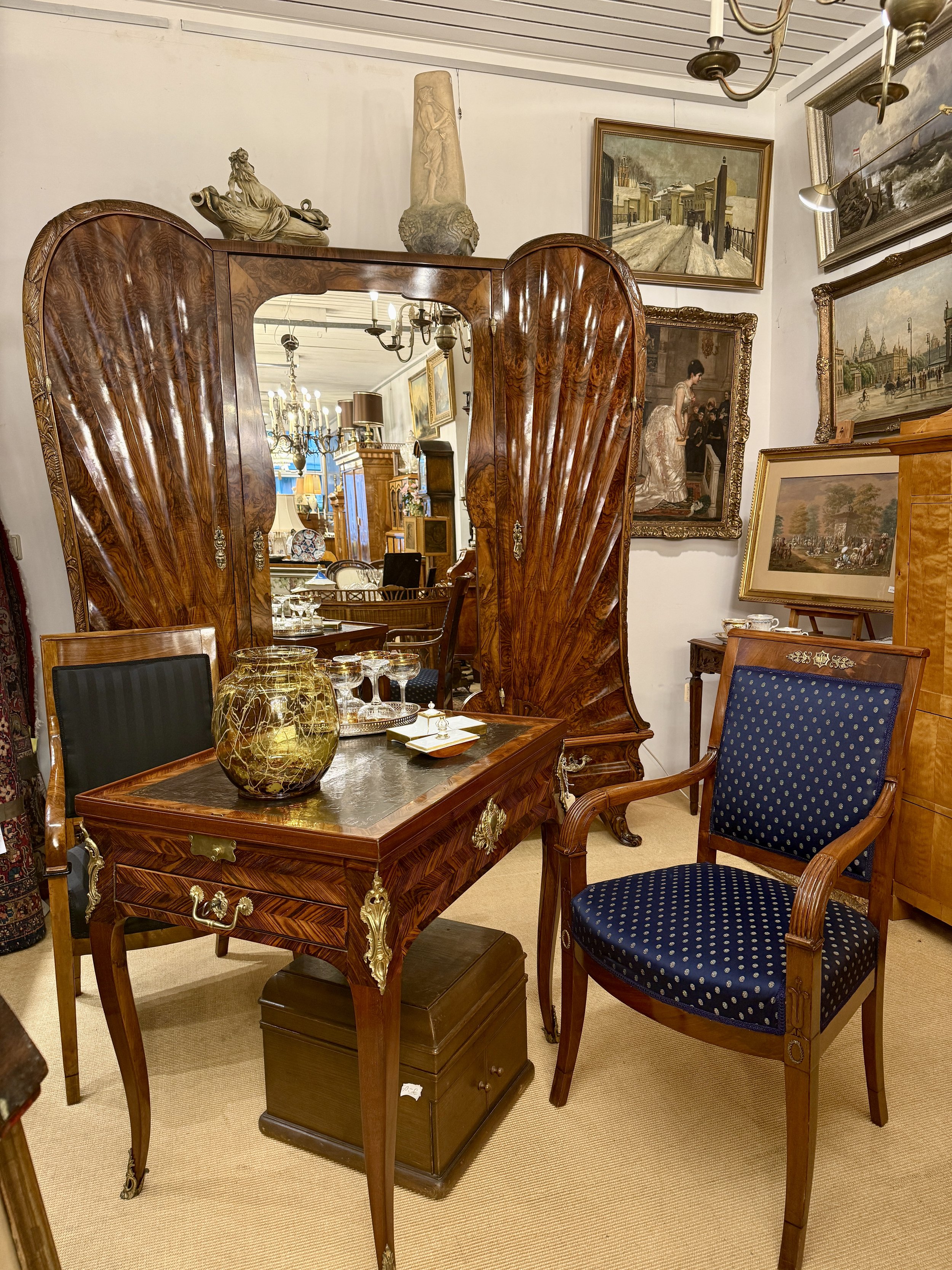 Ein antiker Holztisch mit einer dunkelblauen gepolsterten Stuhllehne und einer geschwungenen Holzarmlehne, dekoriert mit einer großen bunten Vase, in einem Raum mit vielen Gemälden und antiken Möbeln.